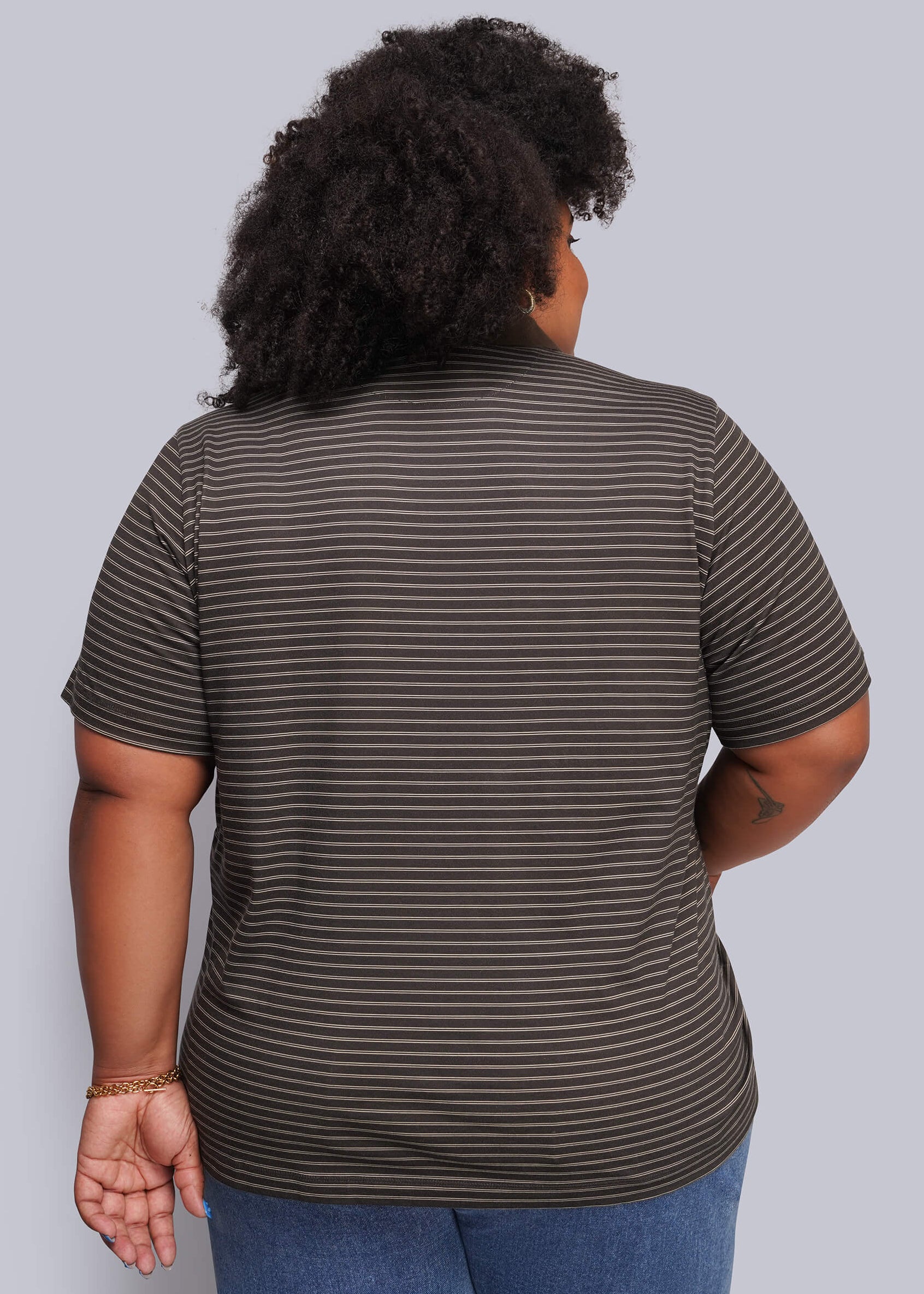 1X=A person viewed from the back wearing a short-sleeve striped top in dark brown and black, paired with blue jeans, against a plain light gray studio background.