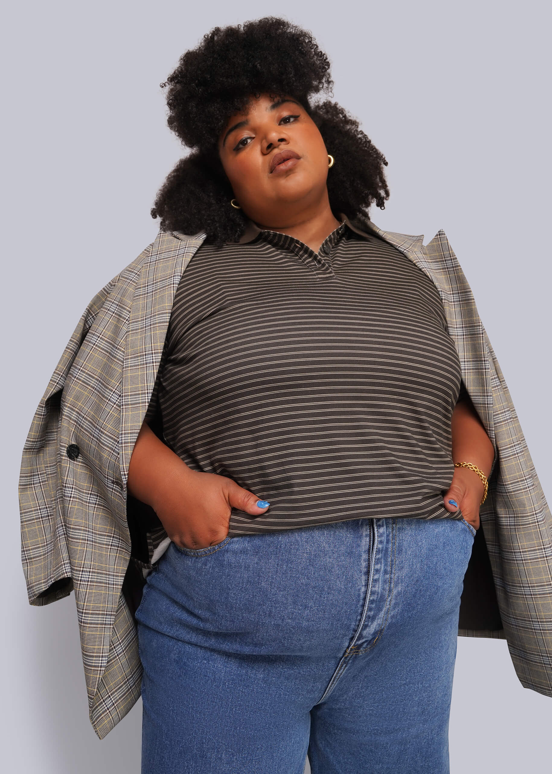 1X=A model wearing a dark brown and gray striped polo shirt, blue jeans, and an open beige plaid blazer, posed against a solid light gray background.