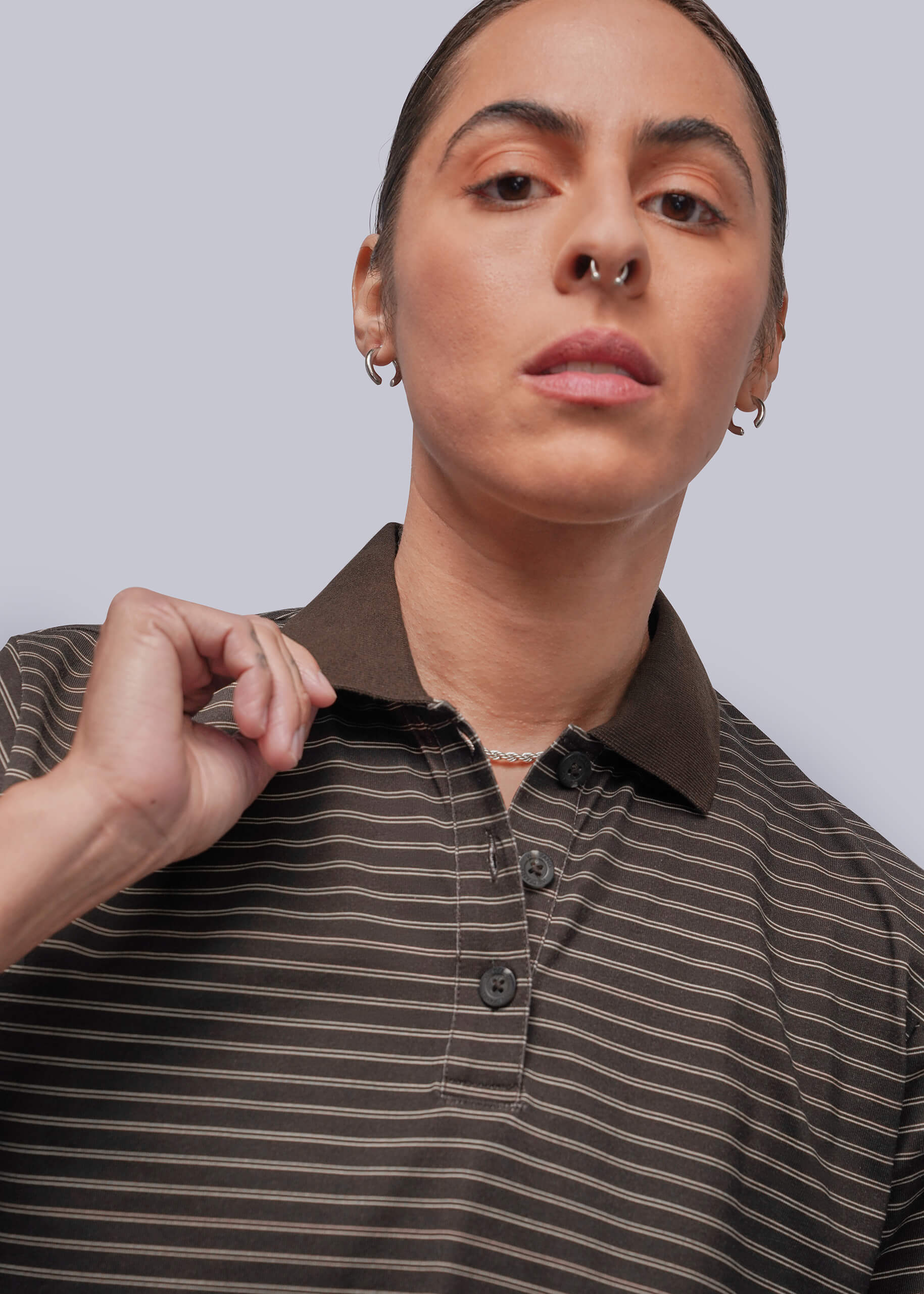 A close-up of a person wearing a brown and dark brown striped polo shirt against a light gray studio background.