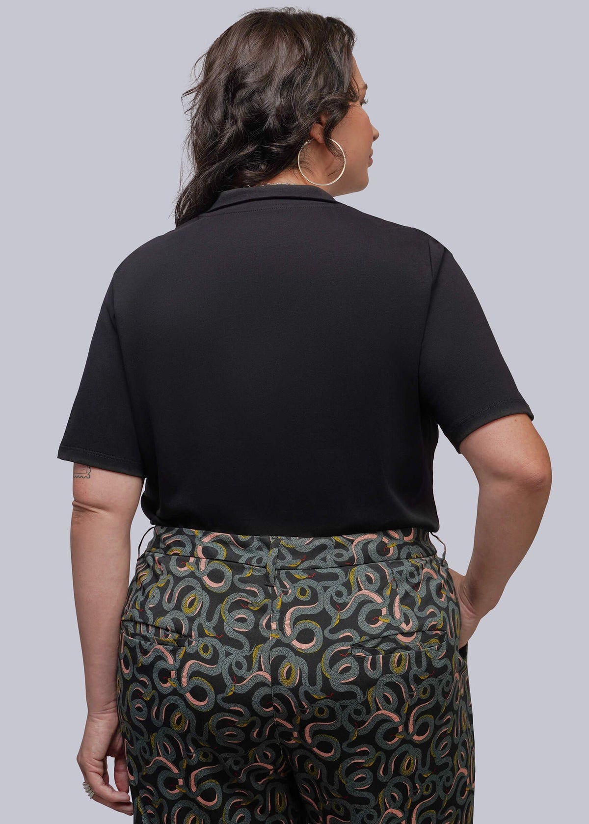 1X=A woman shown from the back wearing a black short-sleeve polo shirt and dark green patterned trousers.