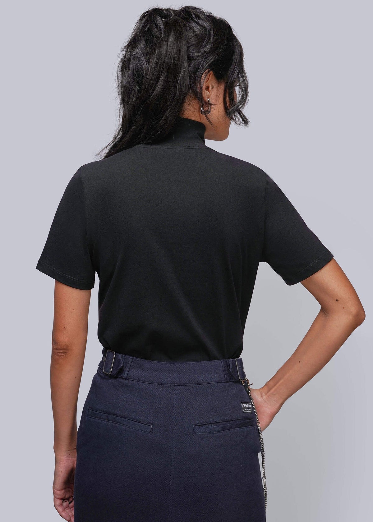 S=A woman shown from the back wearing a black short-sleeve mock neck top and dark navy denim skirt, photographed against a solid light gray studio background.