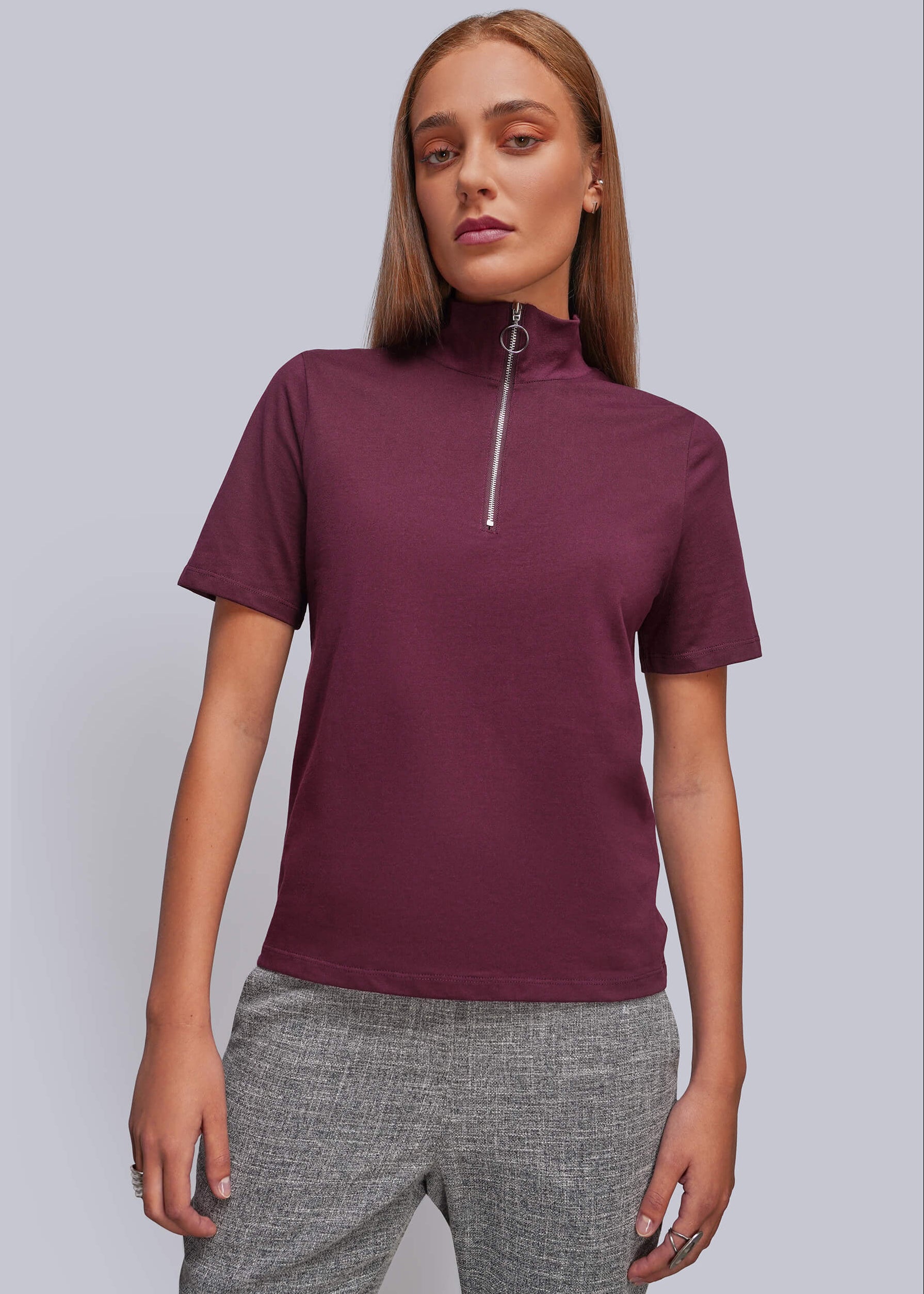 A woman modeling a short-sleeve mock neck top with a silver zipper at the collar, paired with gray textured pants, against a solid light gray background.