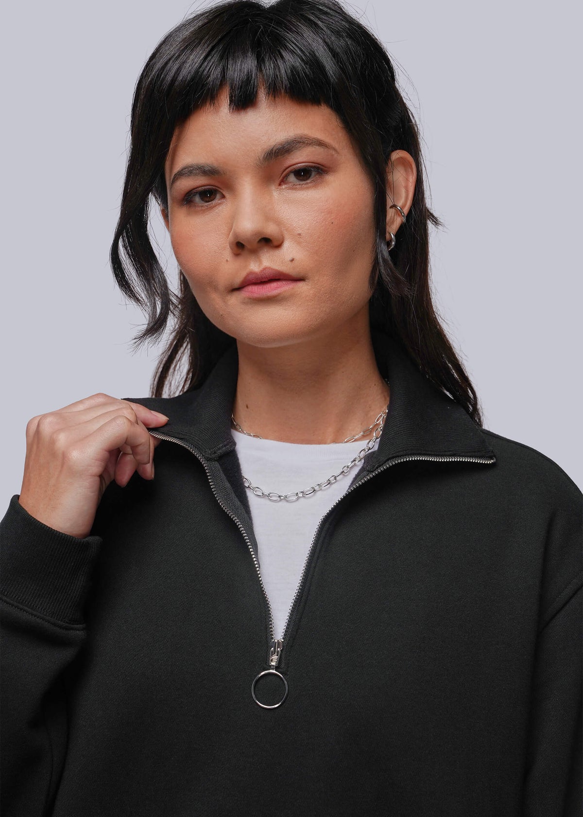 S=A woman with dark hair and bangs wearing a black half-zip sweatshirt over a white t-shirt, accessorized with silver chain necklaces, against a solid gray background.