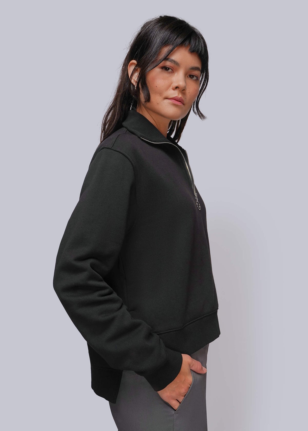 S=A woman wearing a black long-sleeve quarter-zip sweatshirt and gray pants, shown in a side profile against a solid light gray studio background.