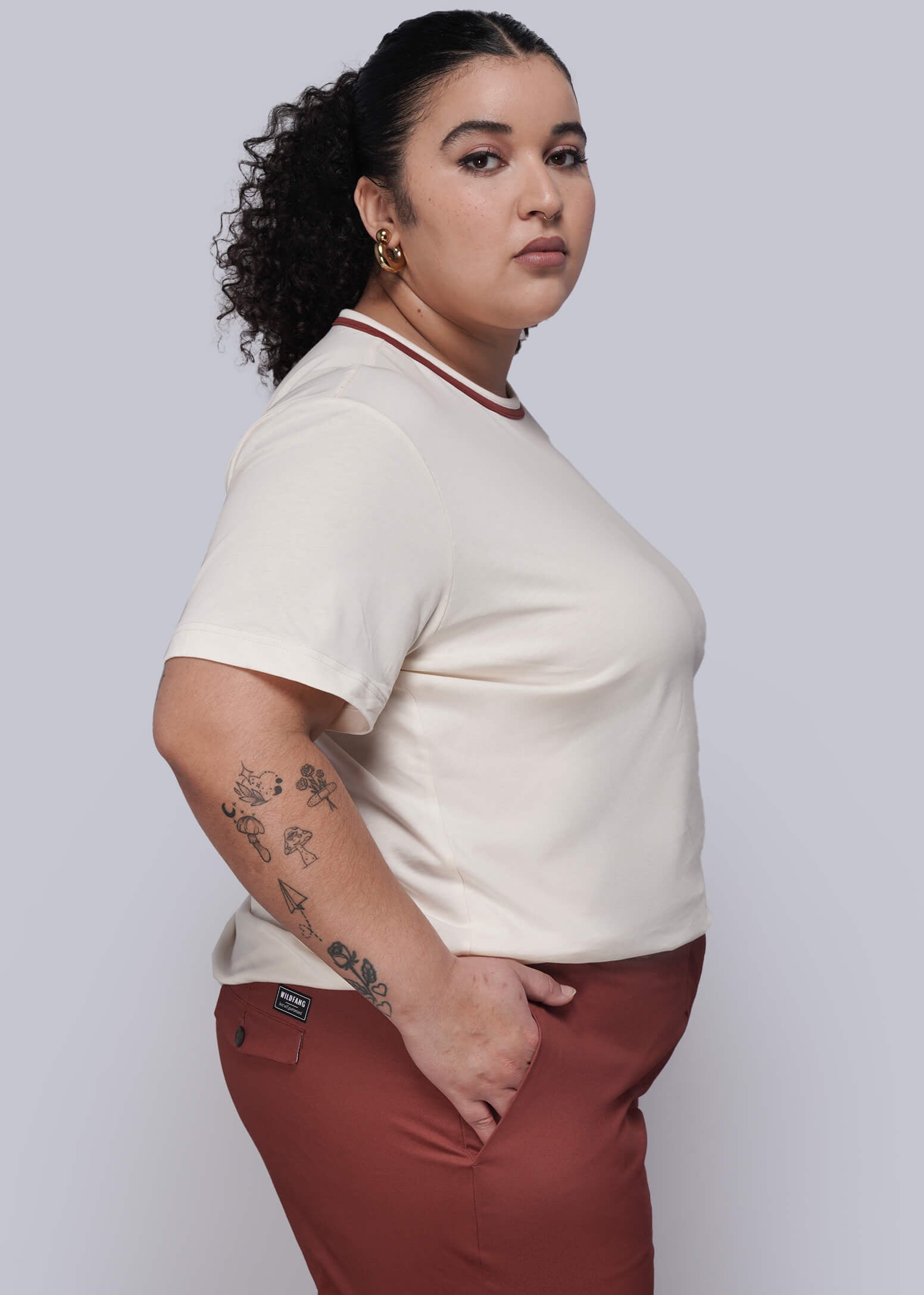 1X=Woman wearing a beige top and rust-colored pants against a white background