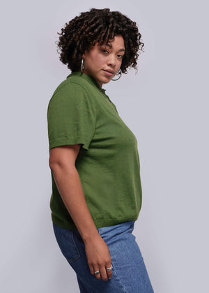 1X=Woman wearing a green polo shirt and blue jeans against a plain background