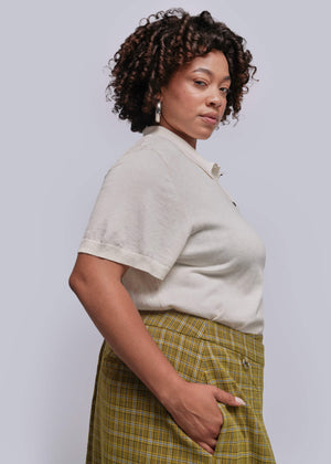 1X=Woman wearing a light-colored shirt and green plaid skirt against a plain background