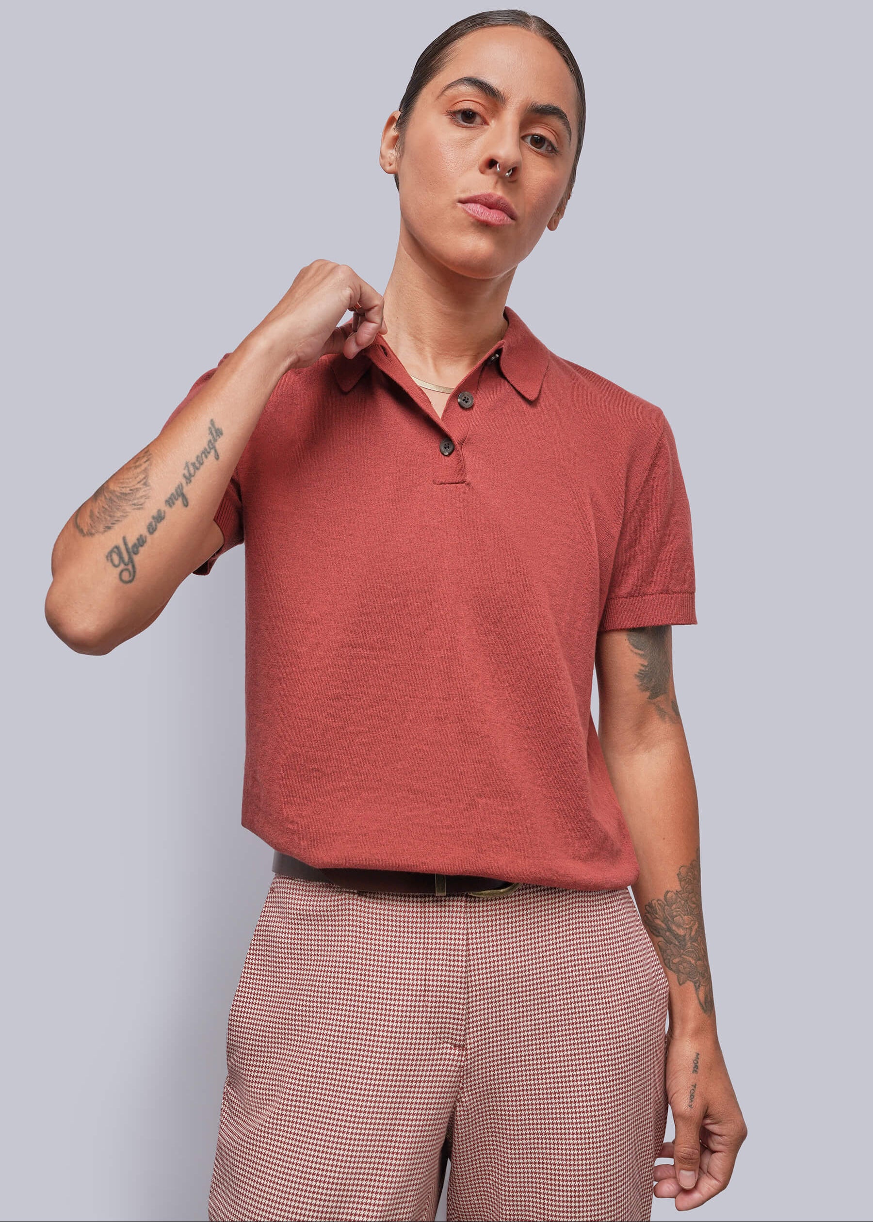 A model wearing a short-sleeve polo shirt in a muted terracotta or dusty rose-red color, paired with red and white houndstooth trousers, against a solid light gray background.