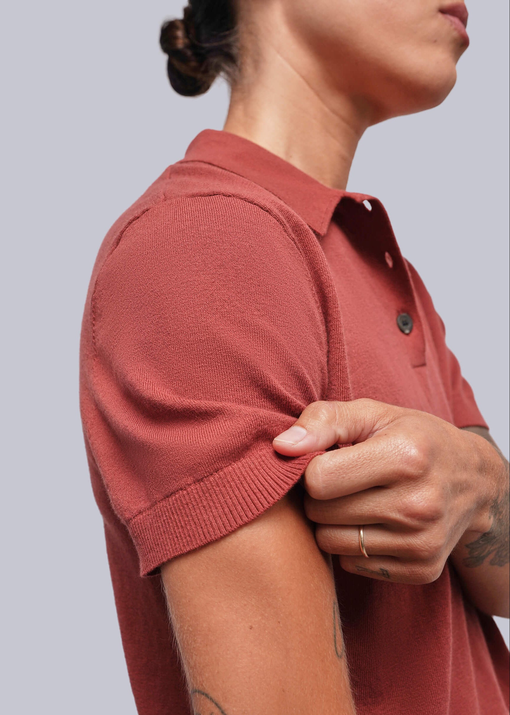 S=Close-up of a person wearing a short-sleeve ribbed knit polo shirt in a muted terracotta or dusty rose color, with the person's hand pulling the sleeve cuff to show the texture.