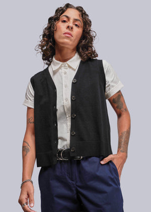 S=Person wearing a black vest over a white shirt with blue pants on a plain background