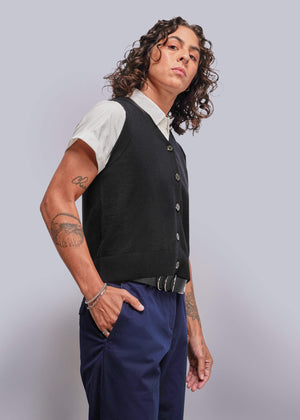 S=Person wearing a black vest over a white shirt with blue pants on a light gray background