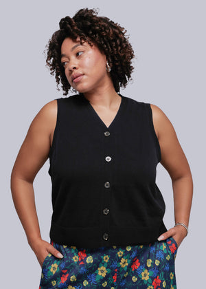 1X=Woman wearing a black sleeveless button-up sweater vest top with colorful floral pants against a gray background