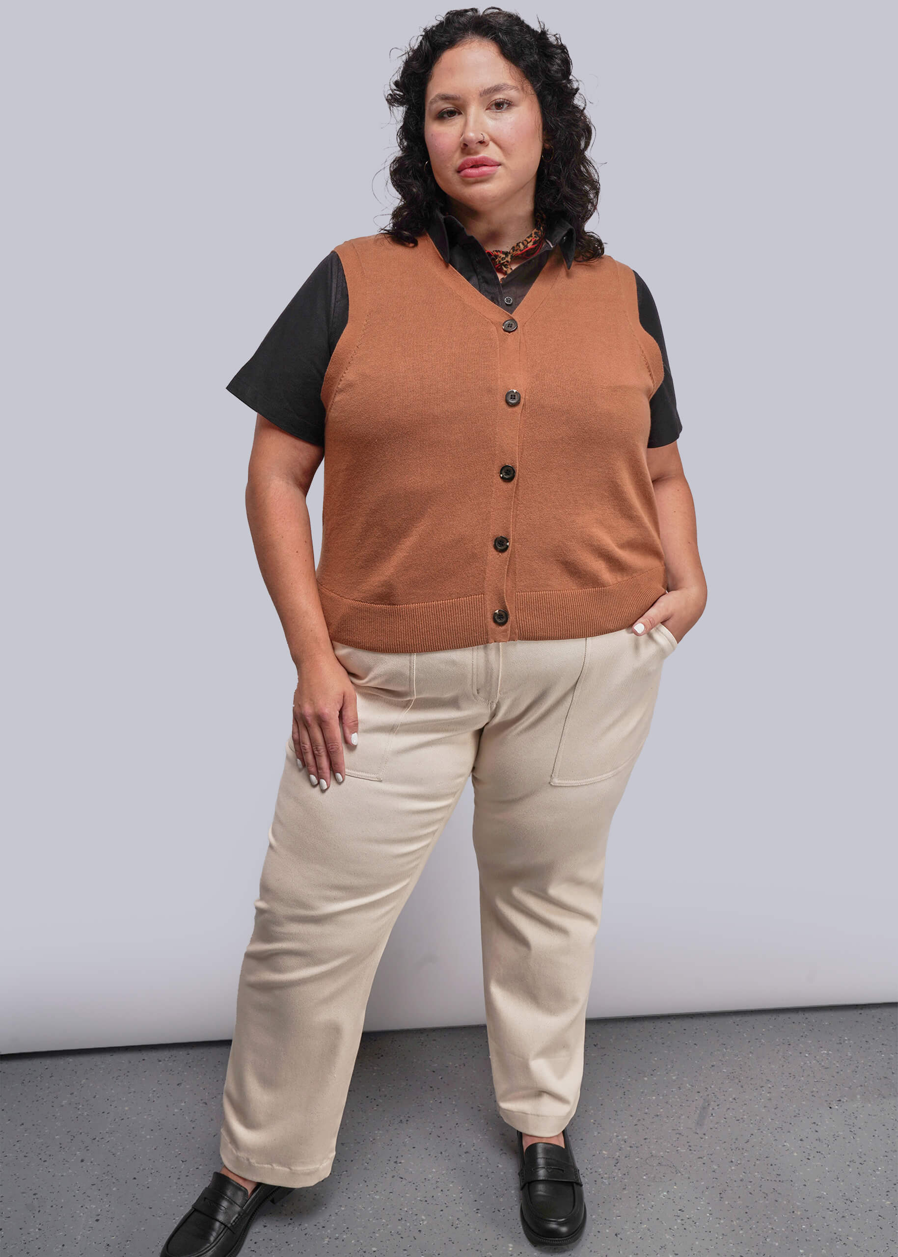1X=Person wearing a brown vest over a black shirt with beige pants against a plain background