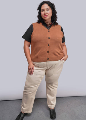 1X=Person wearing a brown vest over a black shirt with beige pants against a plain background