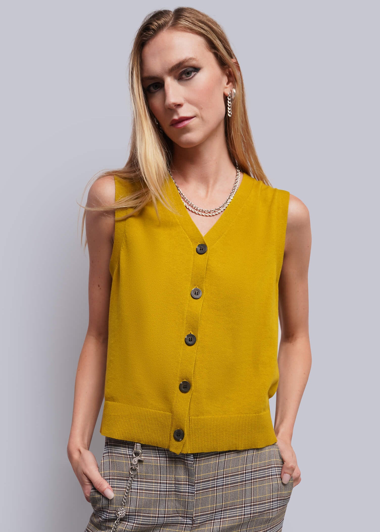 S=A woman modeling a sleeveless mustard yellow knit vest with dark buttons, paired with brown plaid trousers, against a solid gray studio background.