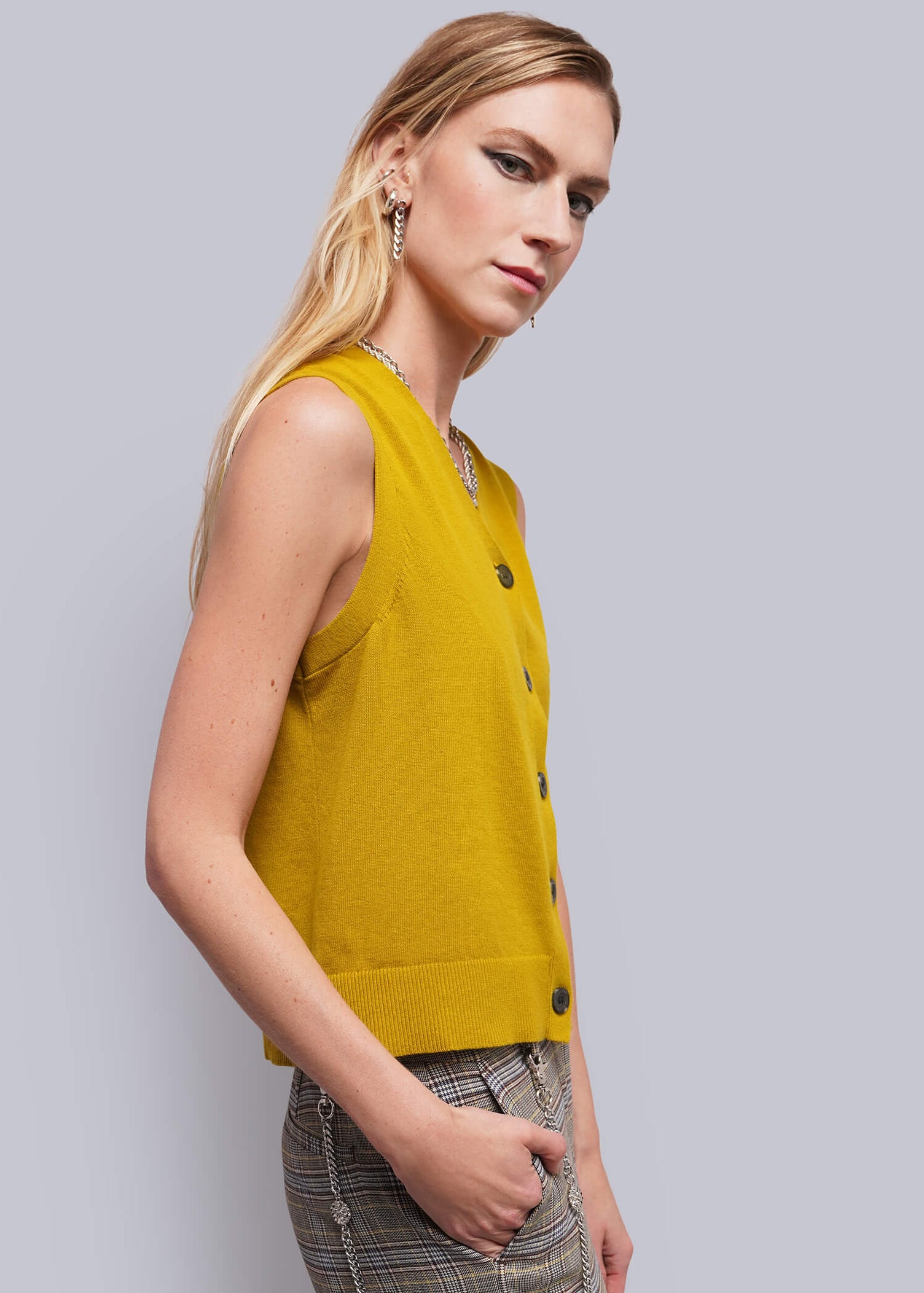 S=A blonde model wearing a sleeveless mustard yellow knit vest with dark buttons and gray plaid trousers, standing against a solid light gray studio background.