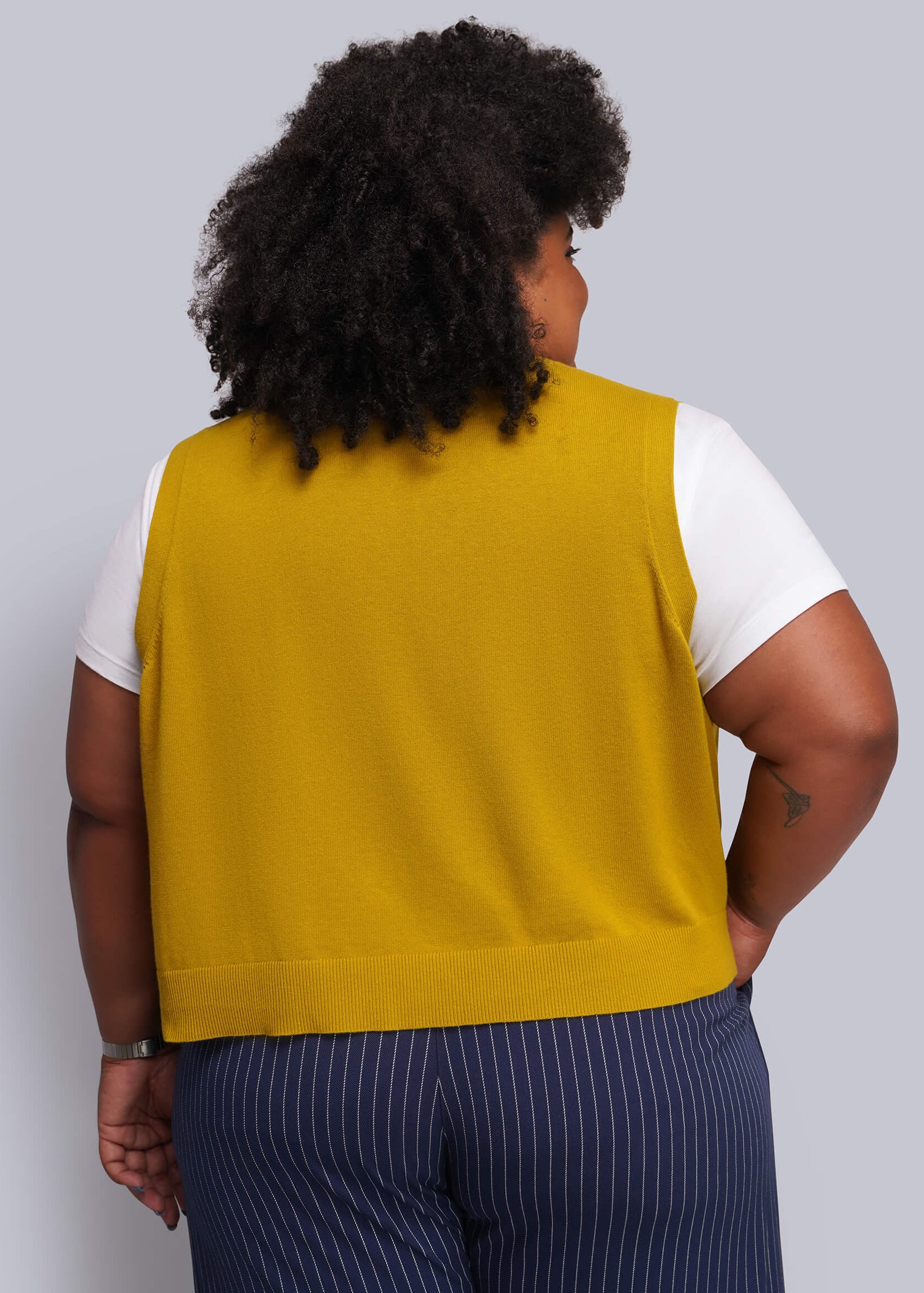 1X=A person shown from the back wearing a sleeveless mustard yellow knit vest over a white short-sleeve top and navy pinstripe pants, against a light gray studio background.