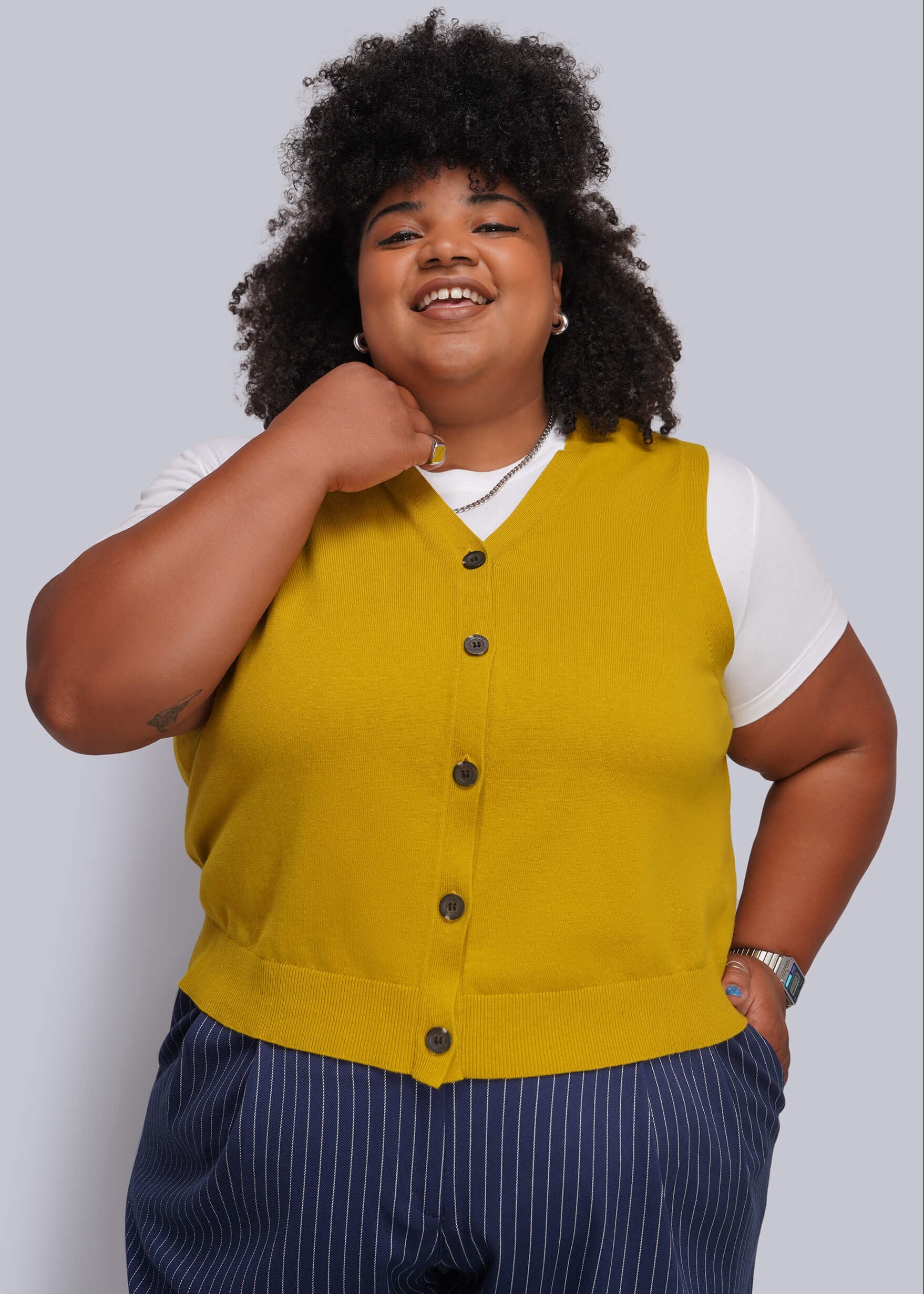 1X=A plus-size model wearing a mustard yellow button-up knit vest over a white t-shirt and navy blue pinstripe trousers, posing against a solid gray studio background.