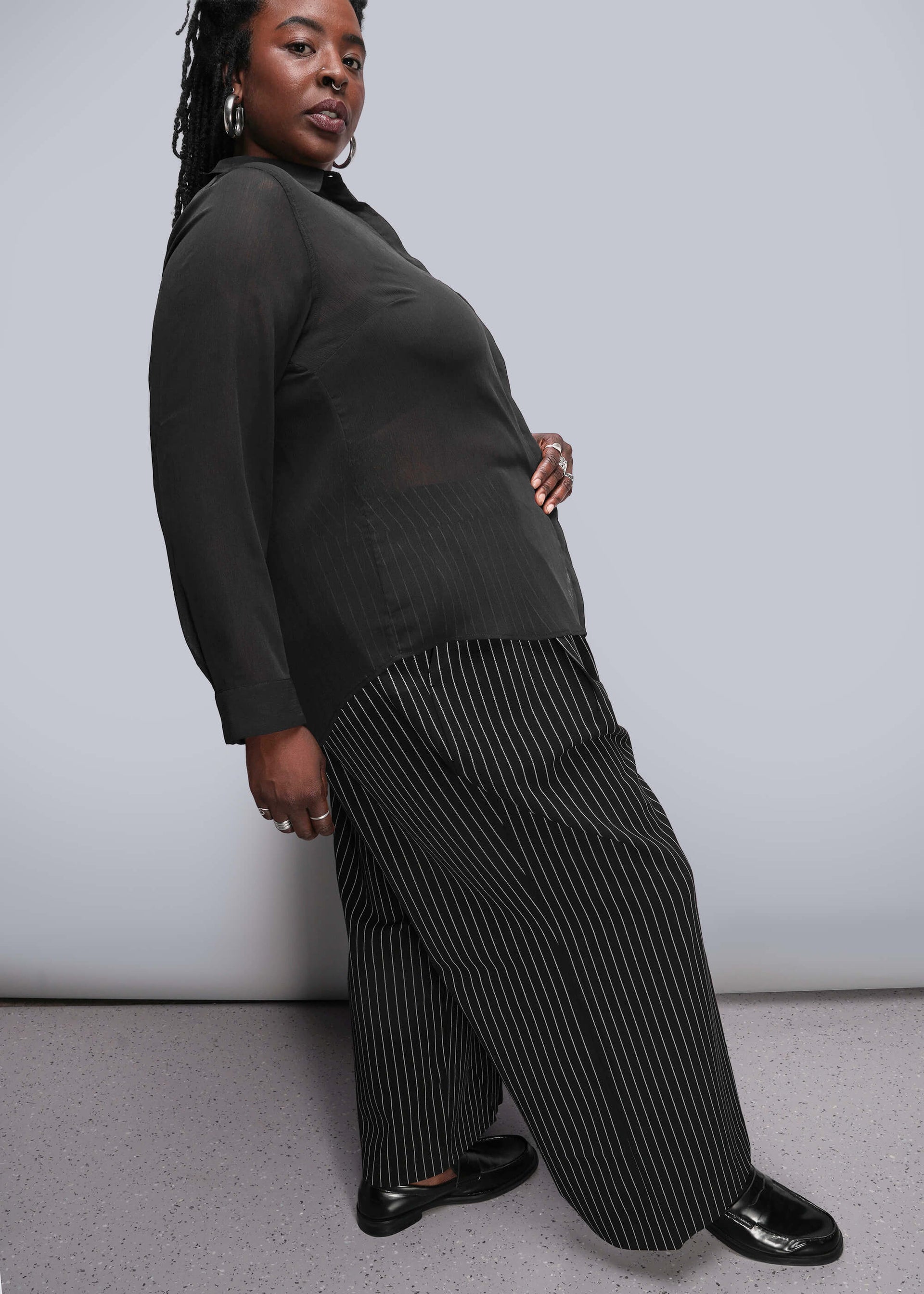 1X=Person wearing a dark sweater and pleated pants against a plain background