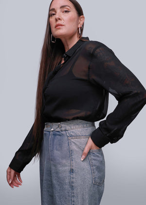 S=Person wearing a black sheer blouse and denim skirt against a gray background