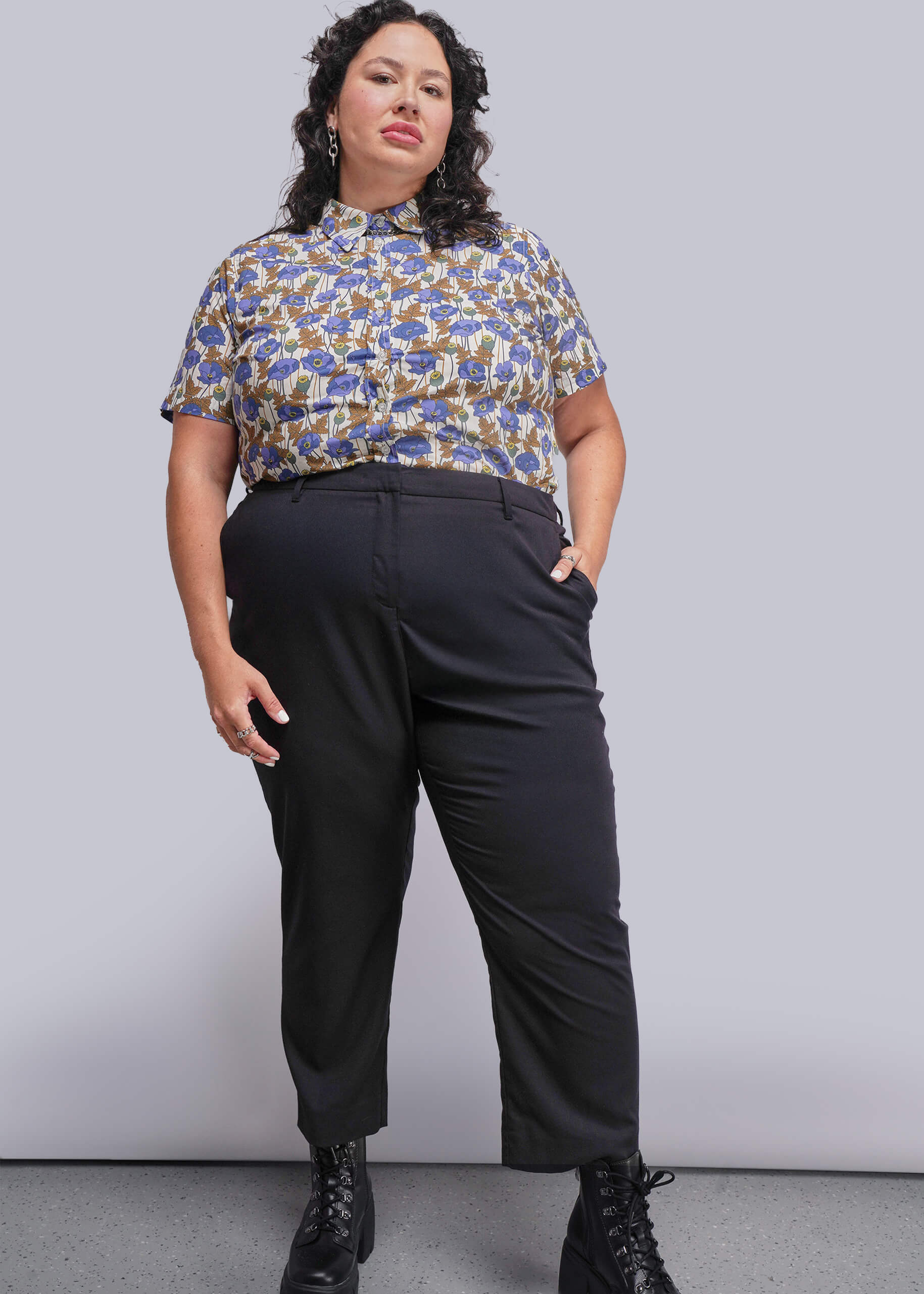 1X=Woman wearing a patterned shirt and black pants against a plain background