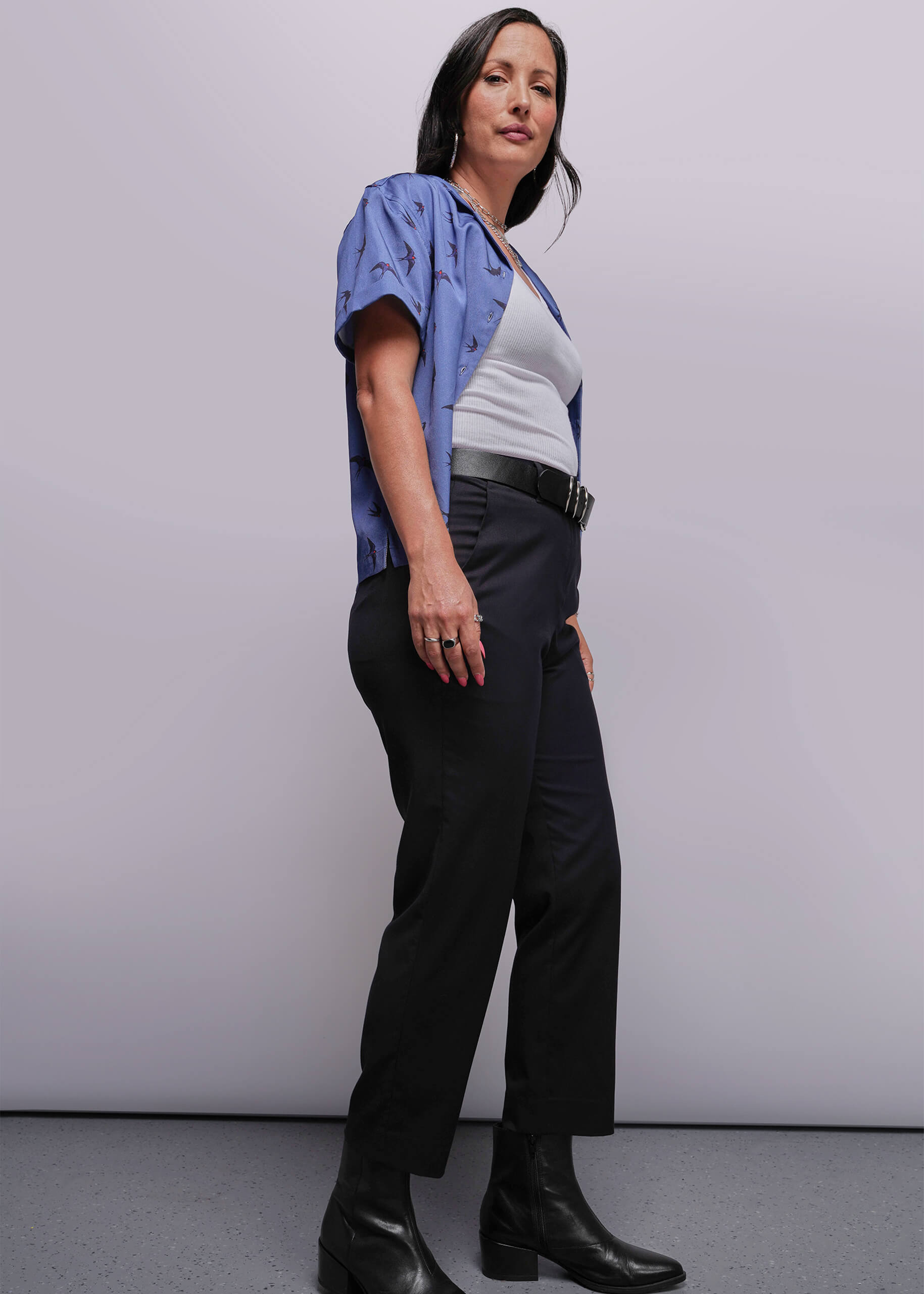 S=Woman wearing a blue shirt, white top, black pants, and black boots against a plain background