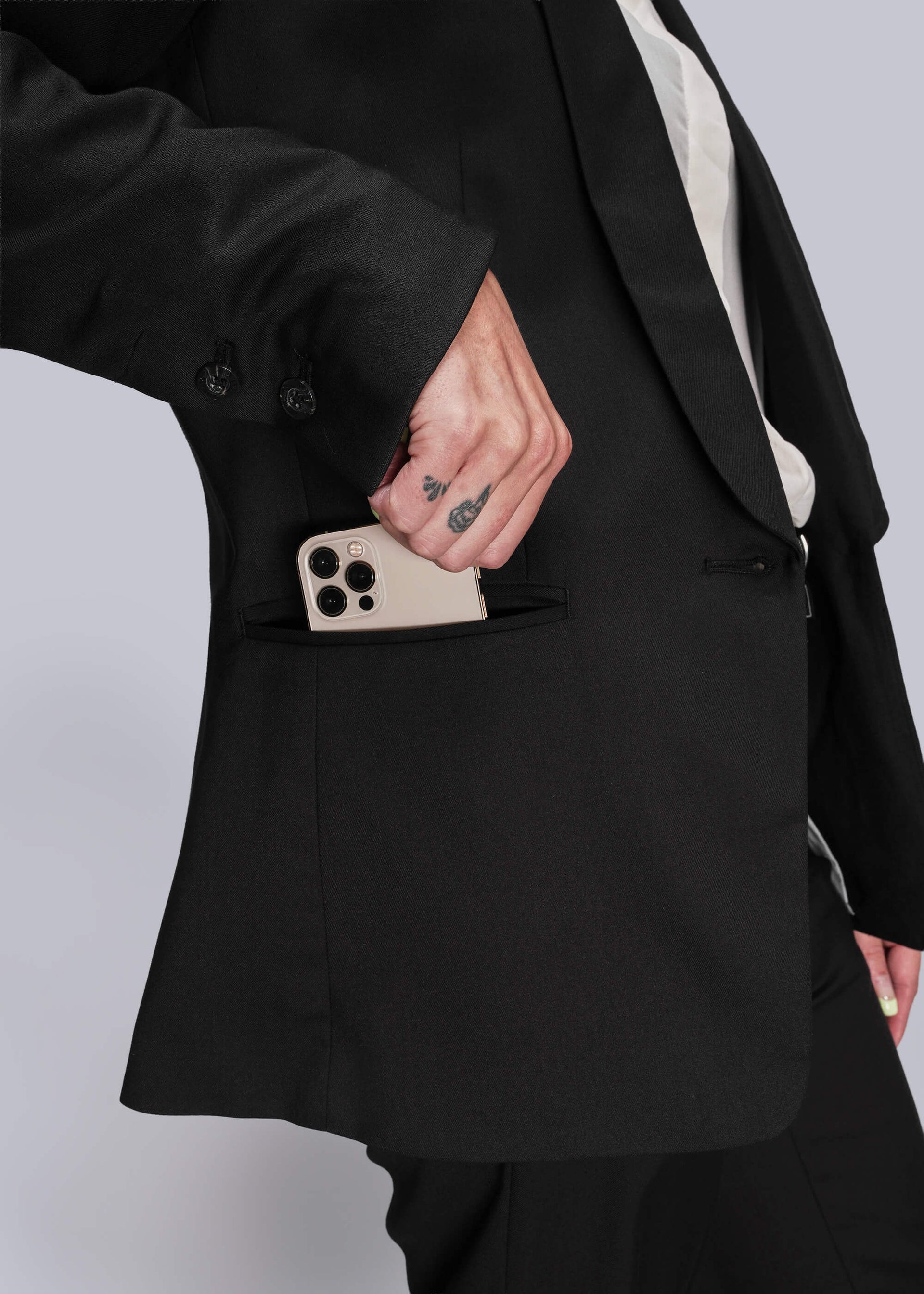 S=Person wearing a black suit with a phone in the pocket against a light gray background