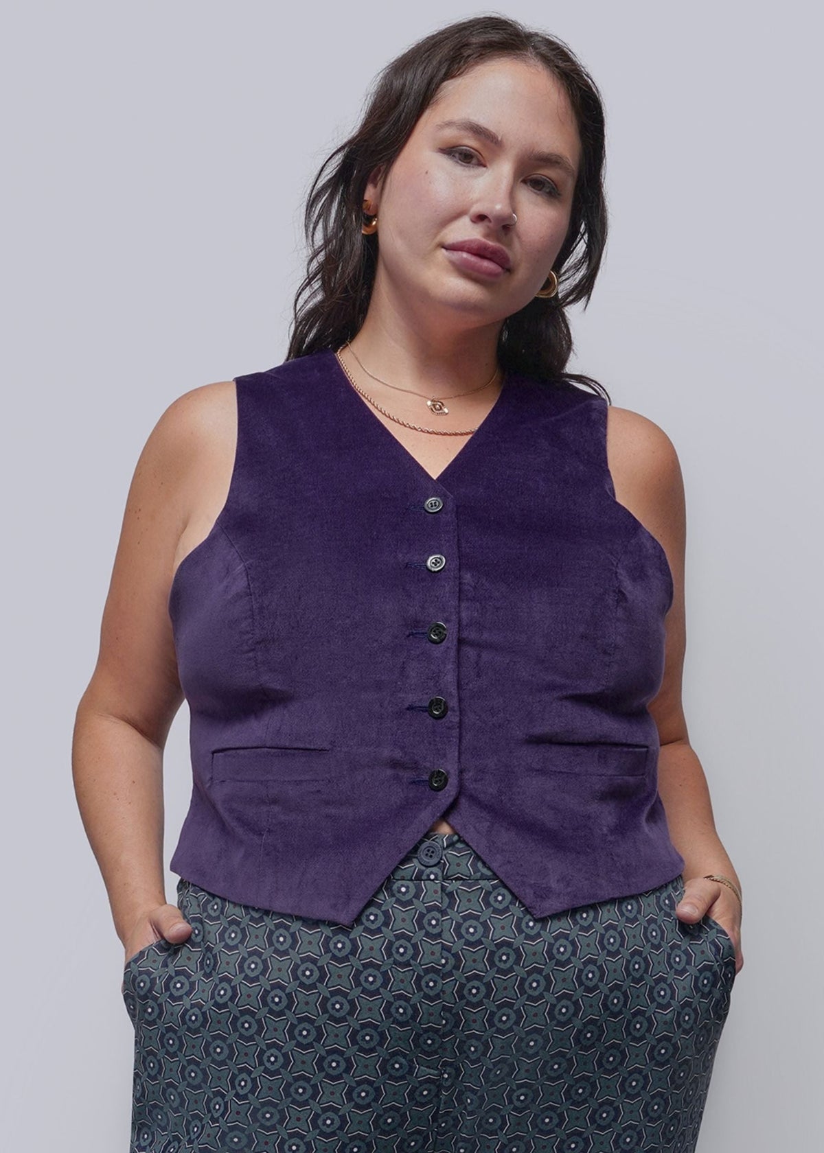 1X=A woman modeling a sleeveless, button-front vest in deep purple, paired with patterned pants, against a solid light gray studio background.