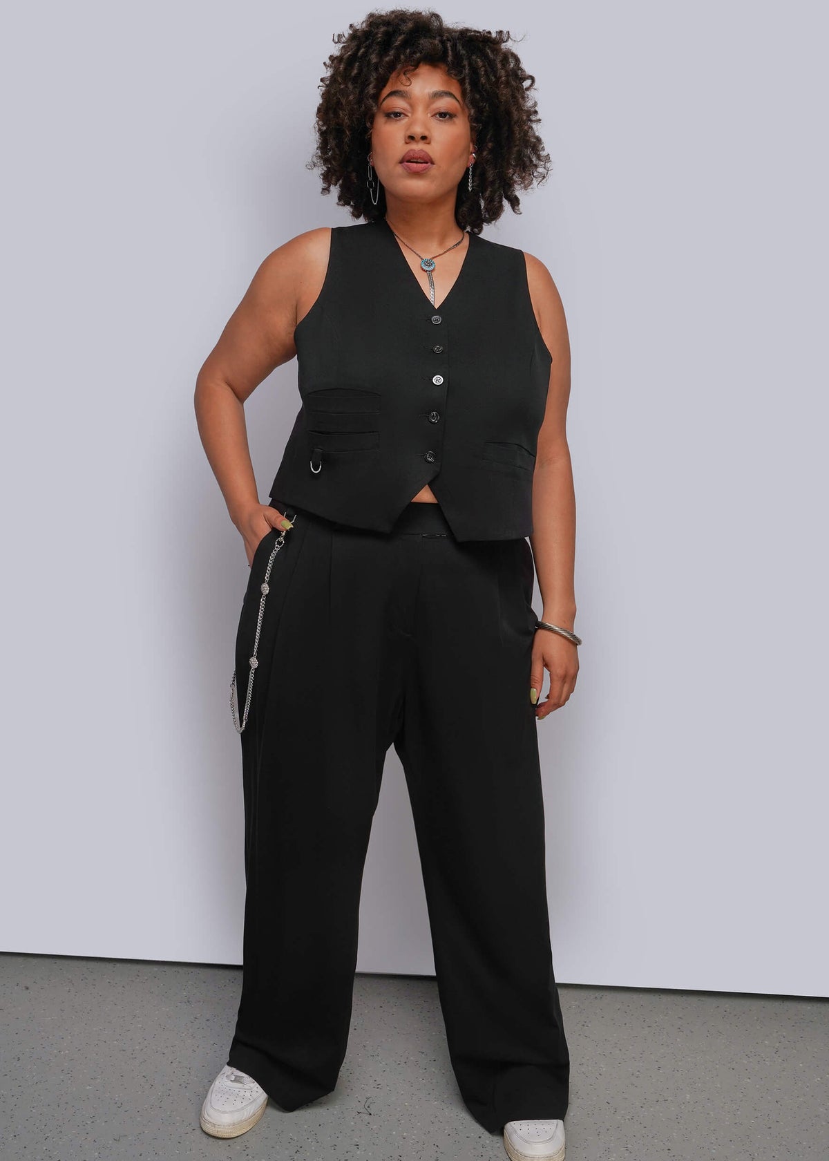 1X=A woman modeling a black sleeveless vest and matching wide-leg trousers with silver zipper details, standing against a light gray studio wall.