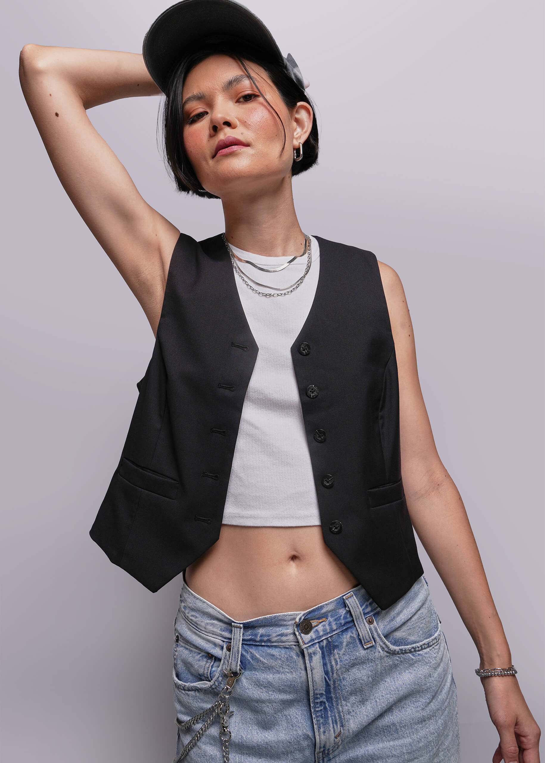 S=Person wearing a black vest over a white shirt and blue jeans against a plain background