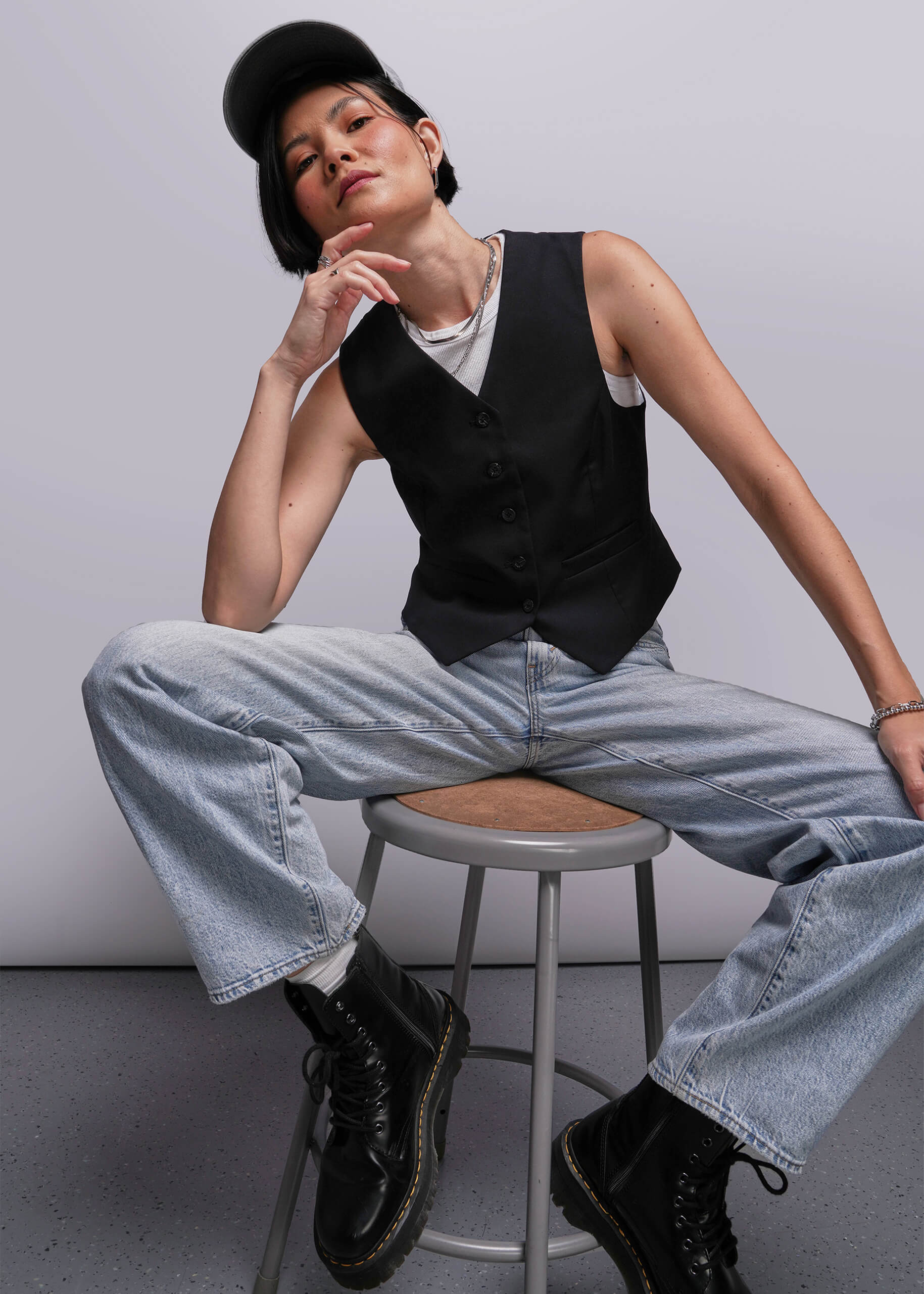 Person sitting on a stool wearing a black vest, white shirt, light blue jeans, and black shoes.
