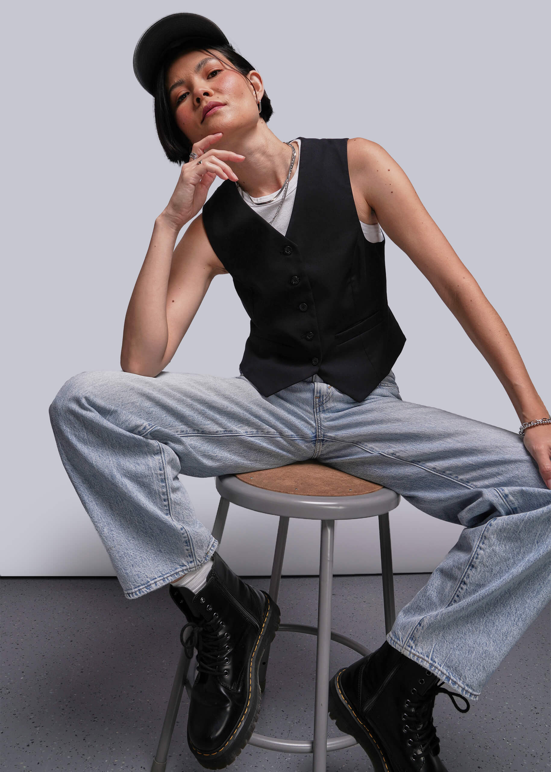Person sitting on a stool wearing a black vest, white shirt, light blue jeans, and black shoes.