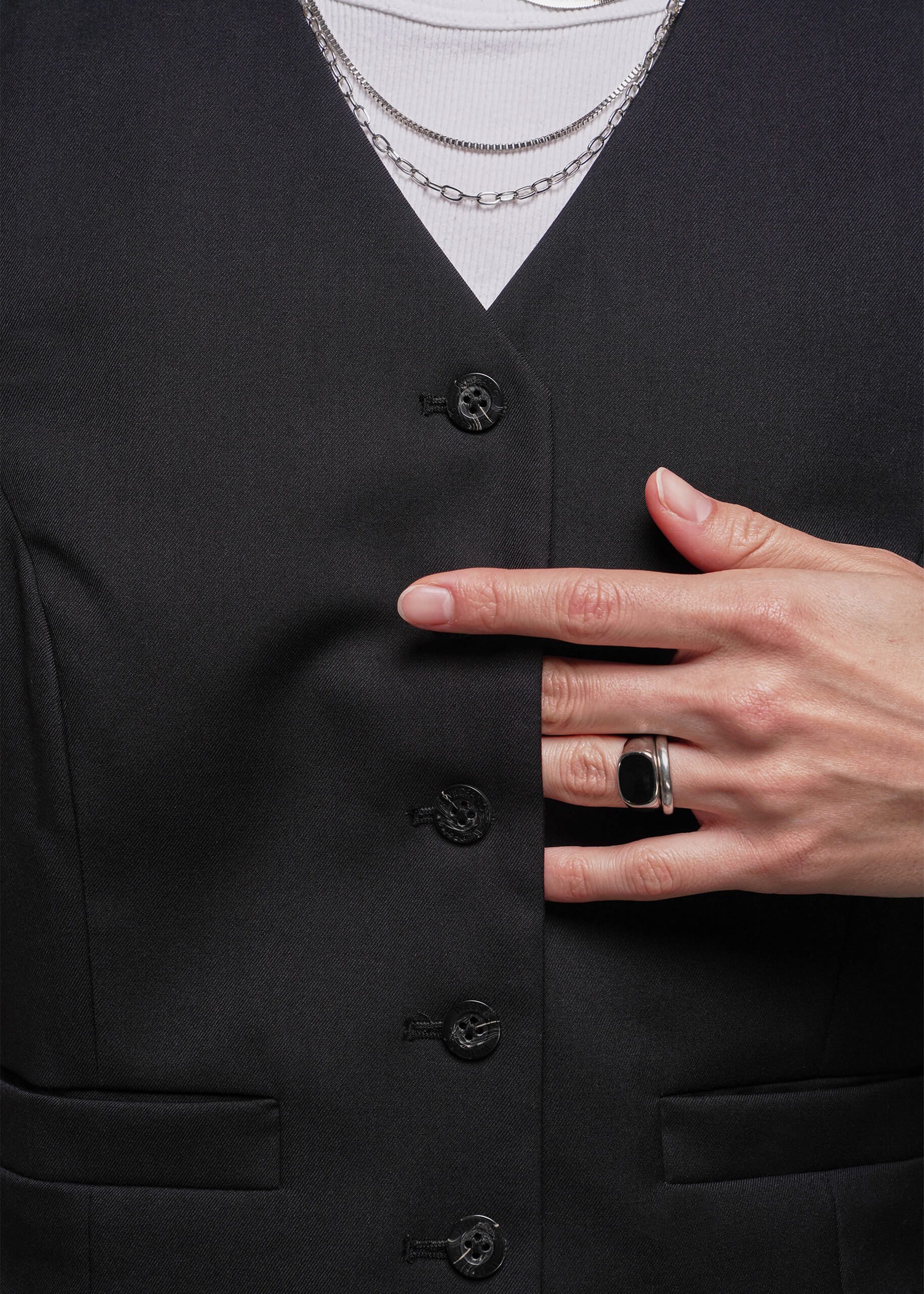 Close up of person wearing a black vest with buttons, white shirt, and layered necklaces.