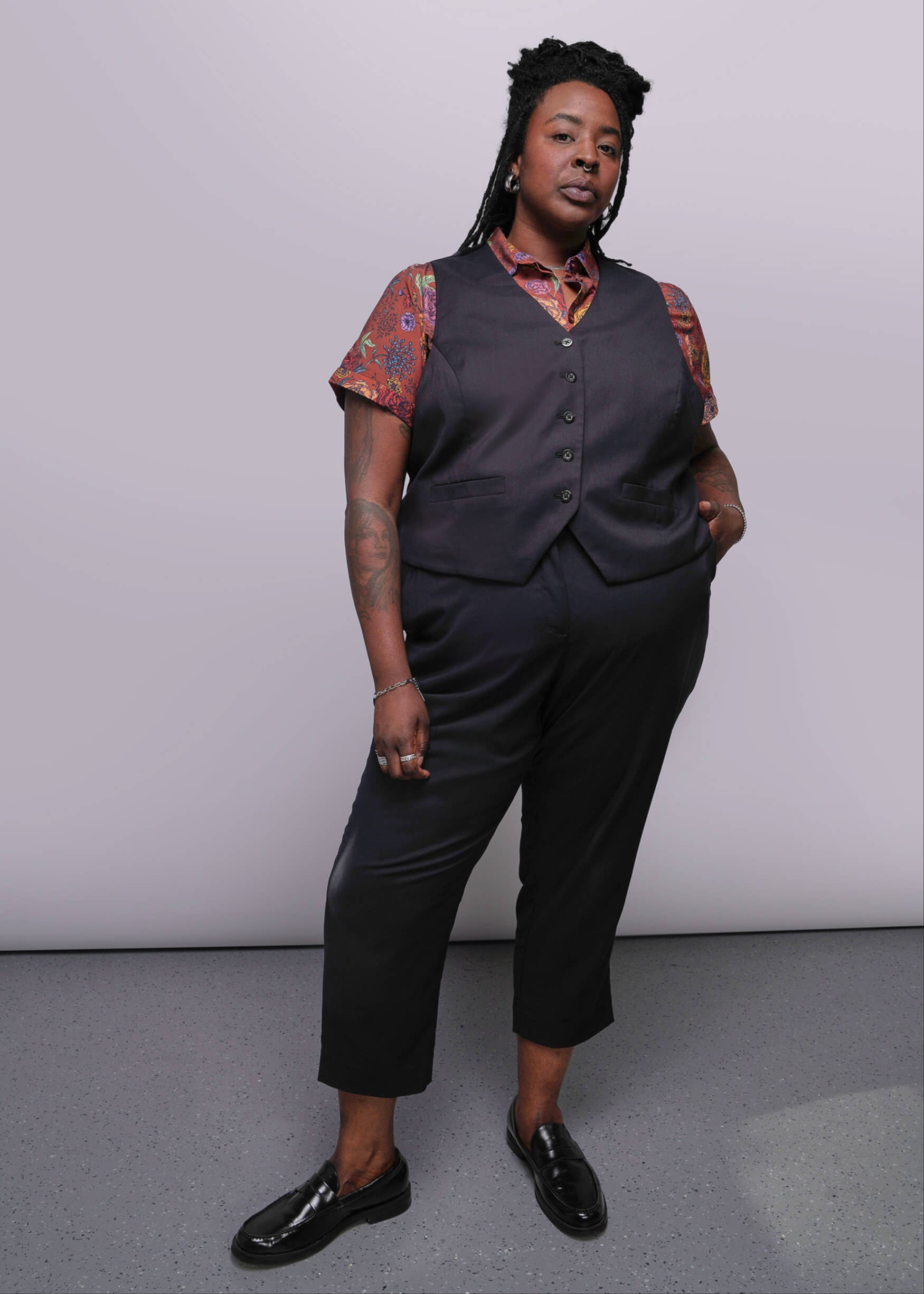 S=Person wearing a dark vest over a patterned shirt with black pants against a plain background