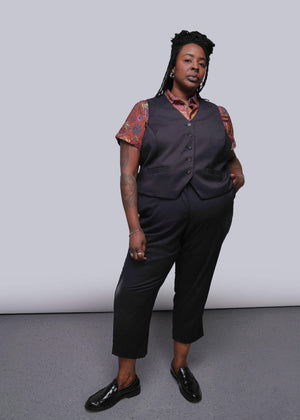 S=Person wearing a dark vest over a patterned shirt with black pants against a plain background