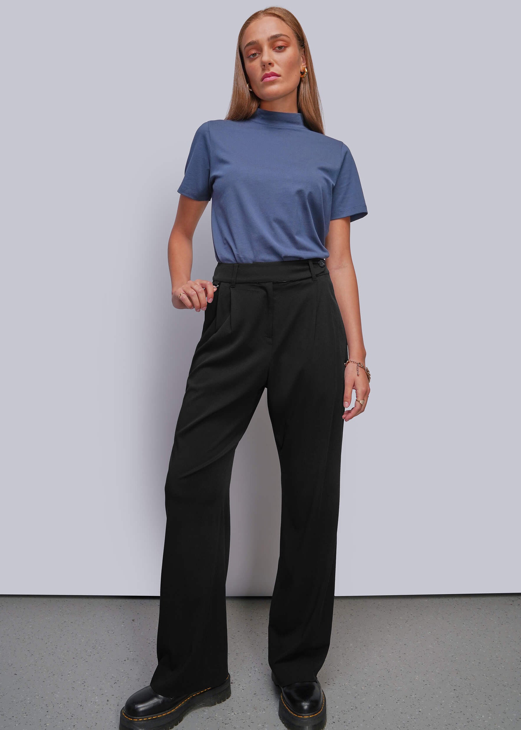 A woman modeling a blue short-sleeve mock neck top and black wide-leg trousers with black platform shoes, standing against a light gray studio wall.