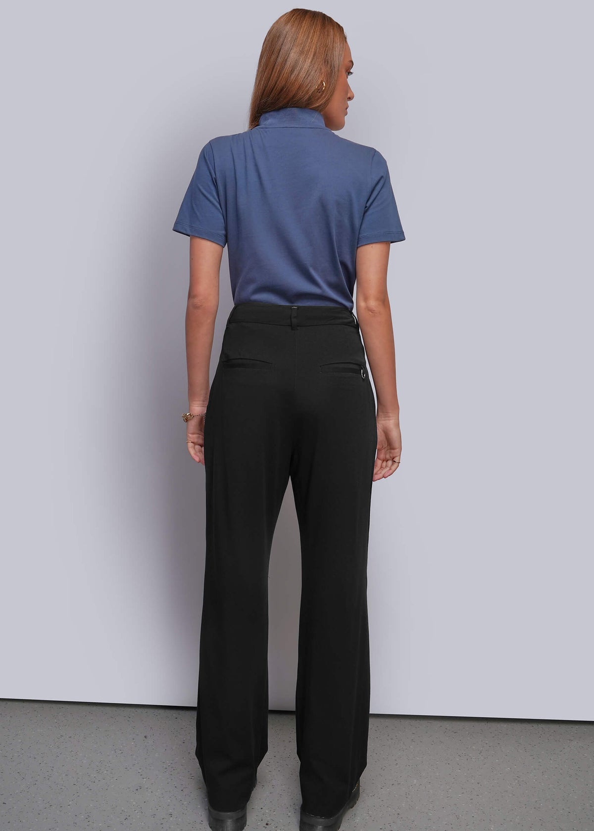S=A woman is shown from the back wearing a slate blue short-sleeve polo shirt and black wide-leg trousers, standing against a light gray wall.