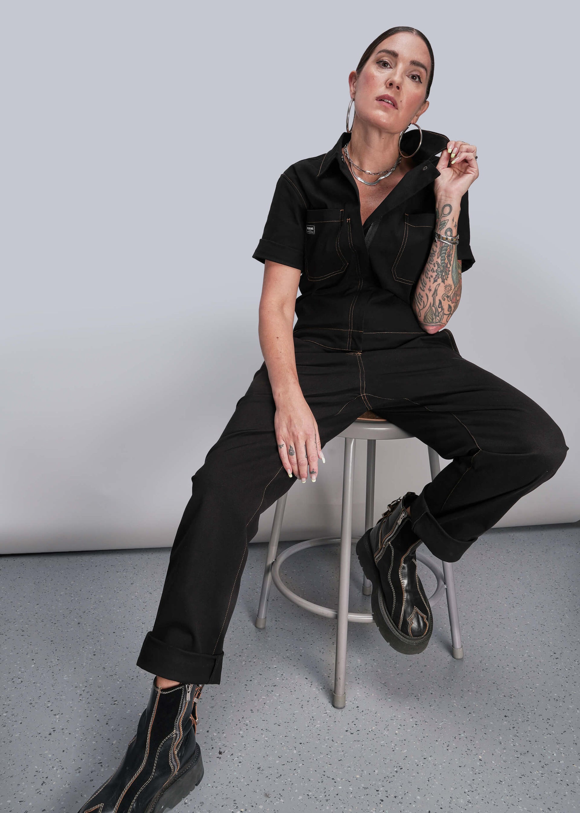 S=Person wearing a black jumpsuit sitting on a stool against a plain background