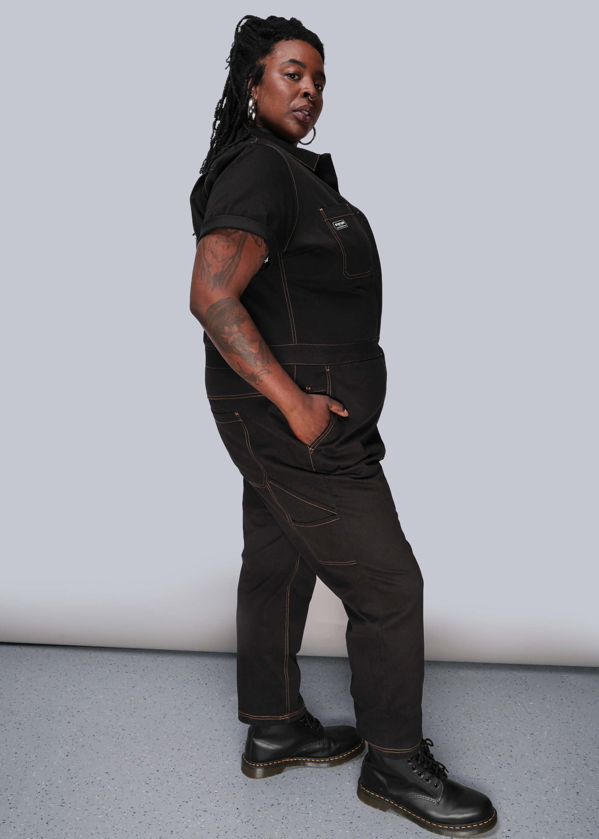 1X=Person wearing a black jumpsuit standing against a plain background