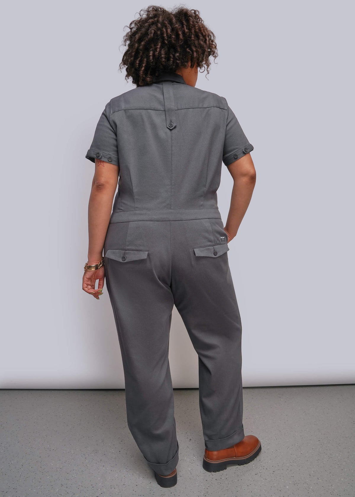 1X=A person shown from the back wearing a gray short-sleeve jumpsuit with flap back pockets and straight legs, standing against a plain light gray wall on a gray floor.