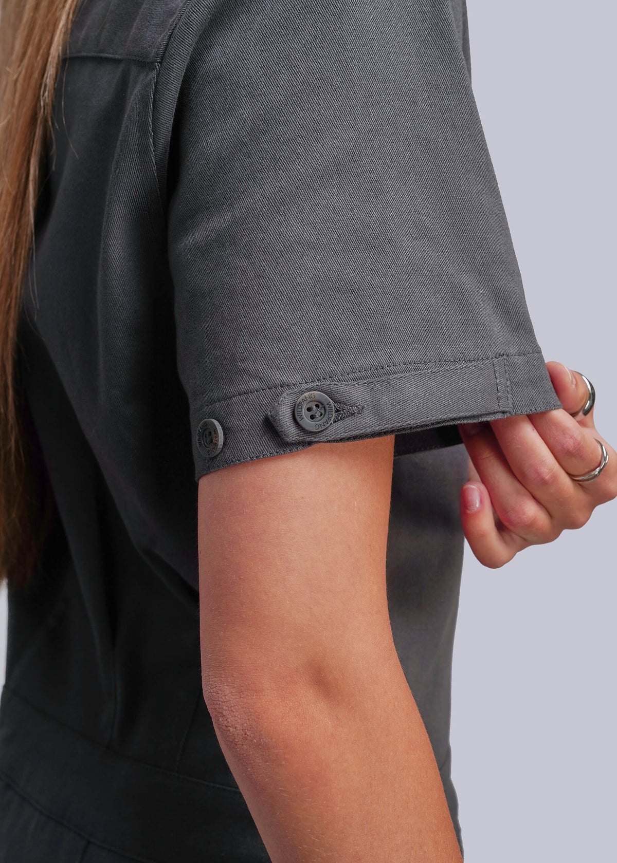 S=Close-up of a person wearing a dark gray short-sleeve jumpsuit, showing the sleeve with two dark buttons and a hand adjusting or holding the cuff against a light gray background.