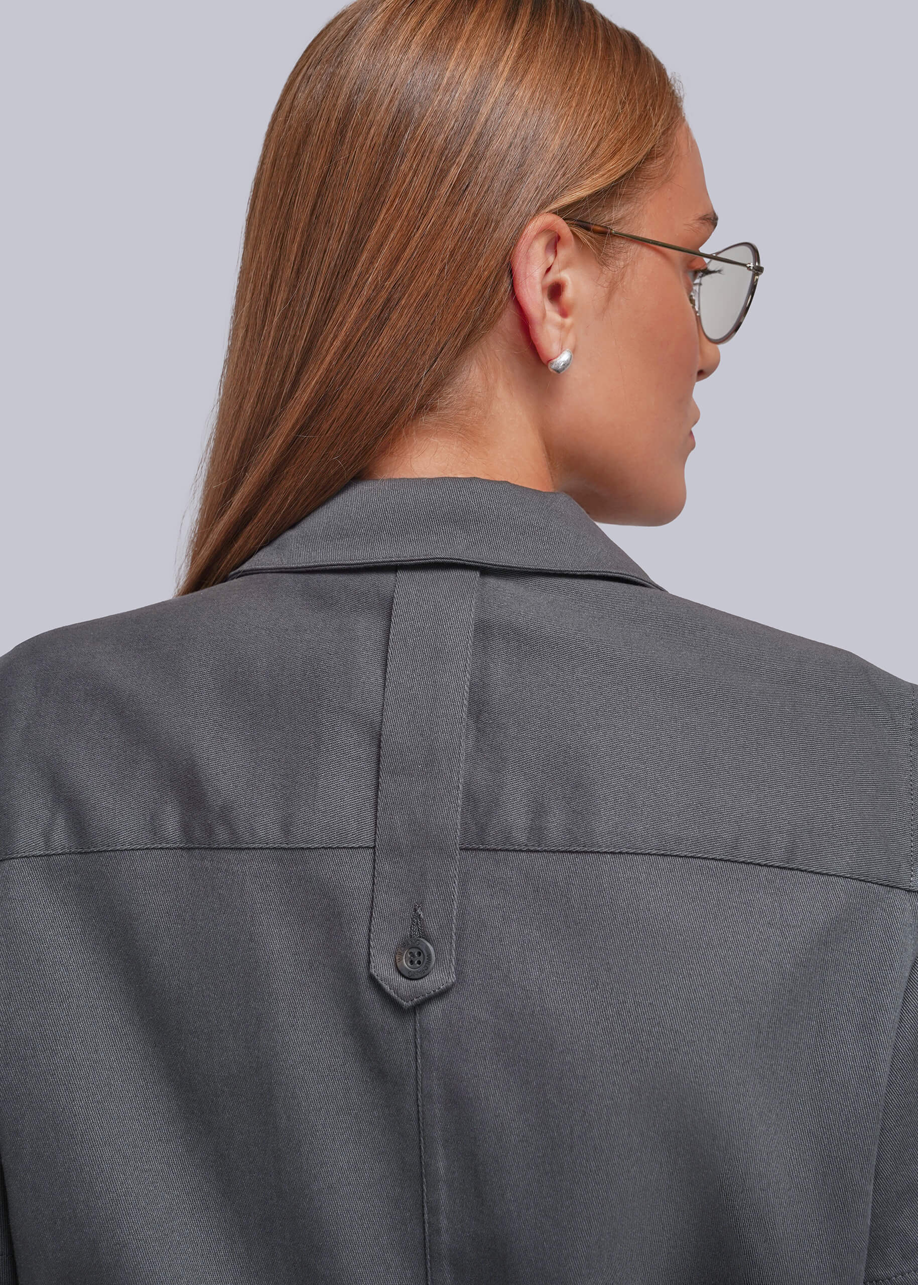 S=Close-up back view of a woman wearing a dark gray collared jumpsuit with a buttoned back detail, shown against a light gray studio background.