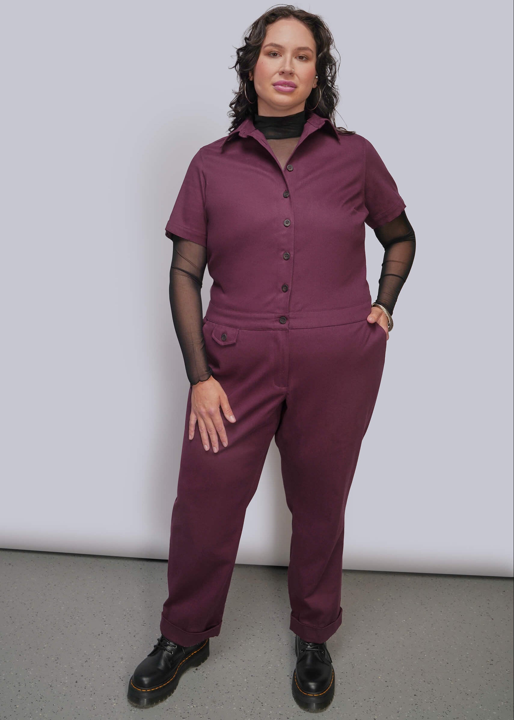 1X=A woman modeling a short sleeve burgundy jumpsuit with black mesh long sleeves, paired with black shoes, against a light gray studio backdrop.