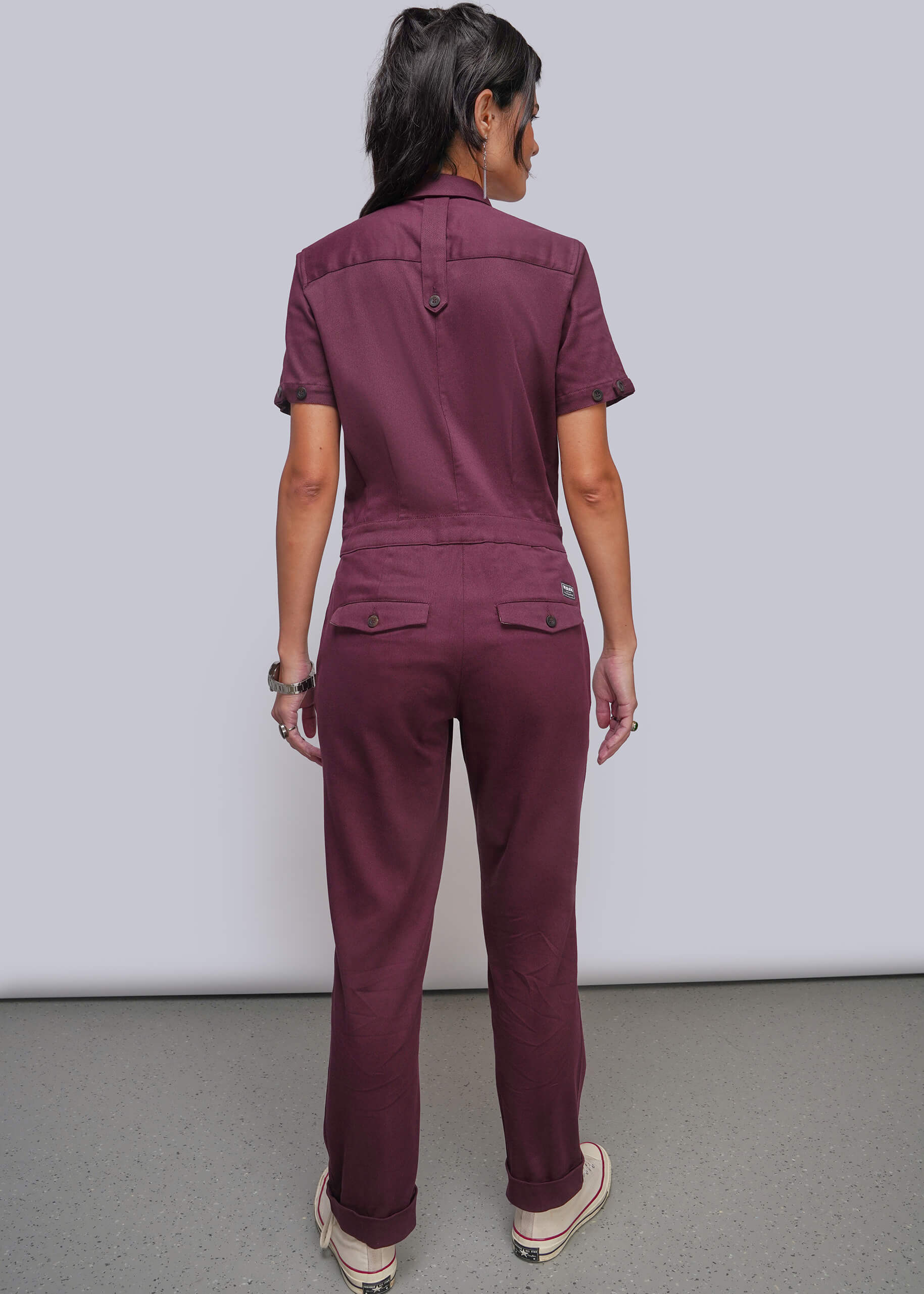 S=A woman viewed from the back wearing a short sleeved jumpsuit in a deep burgundy or plum shade, standing against a plain light gray studio wall.