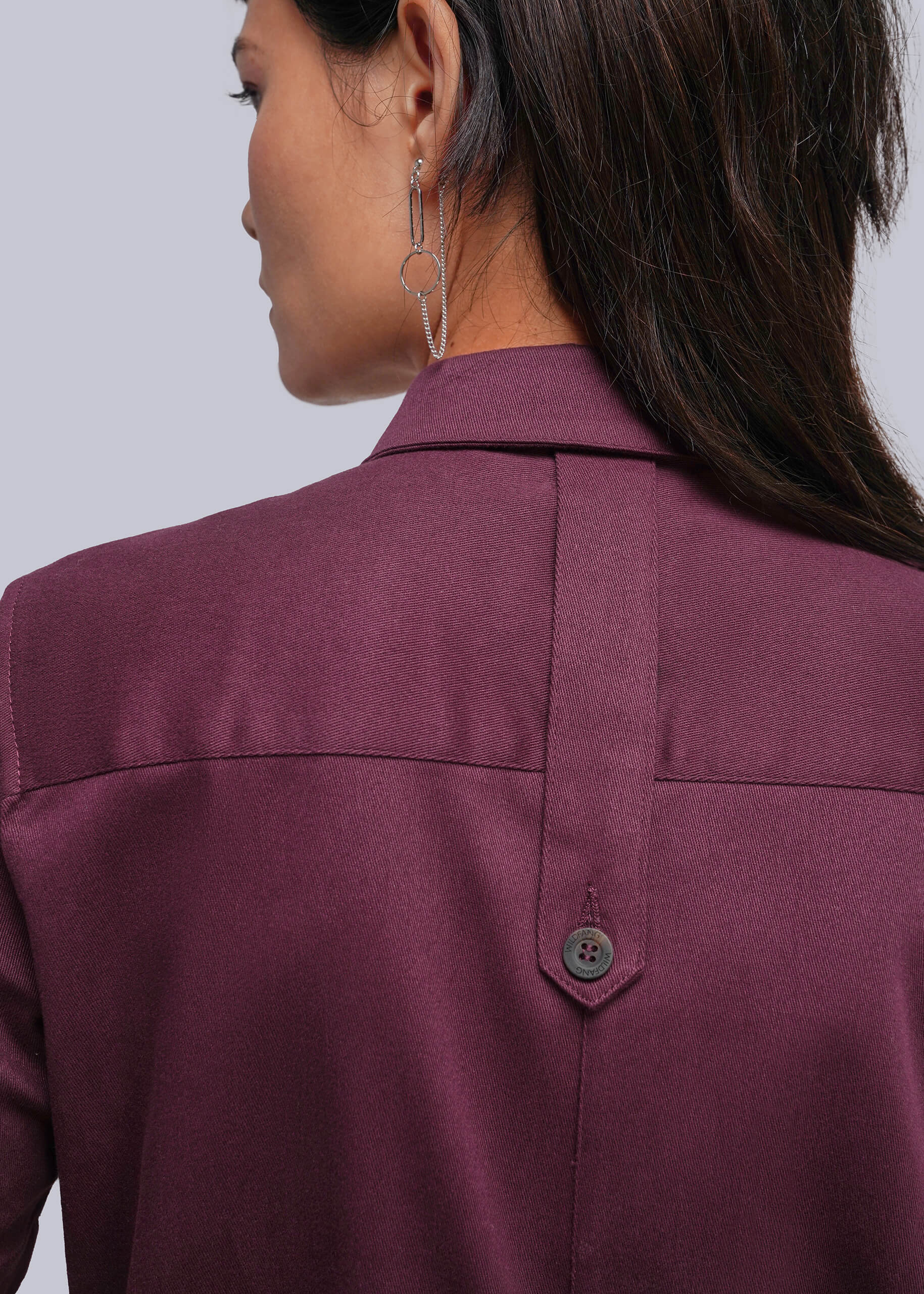 S=Close-up back view of a woman wearing a burgundy jumpsuit, showing the collar, shoulder seams, and a button locker loop detail on the back.