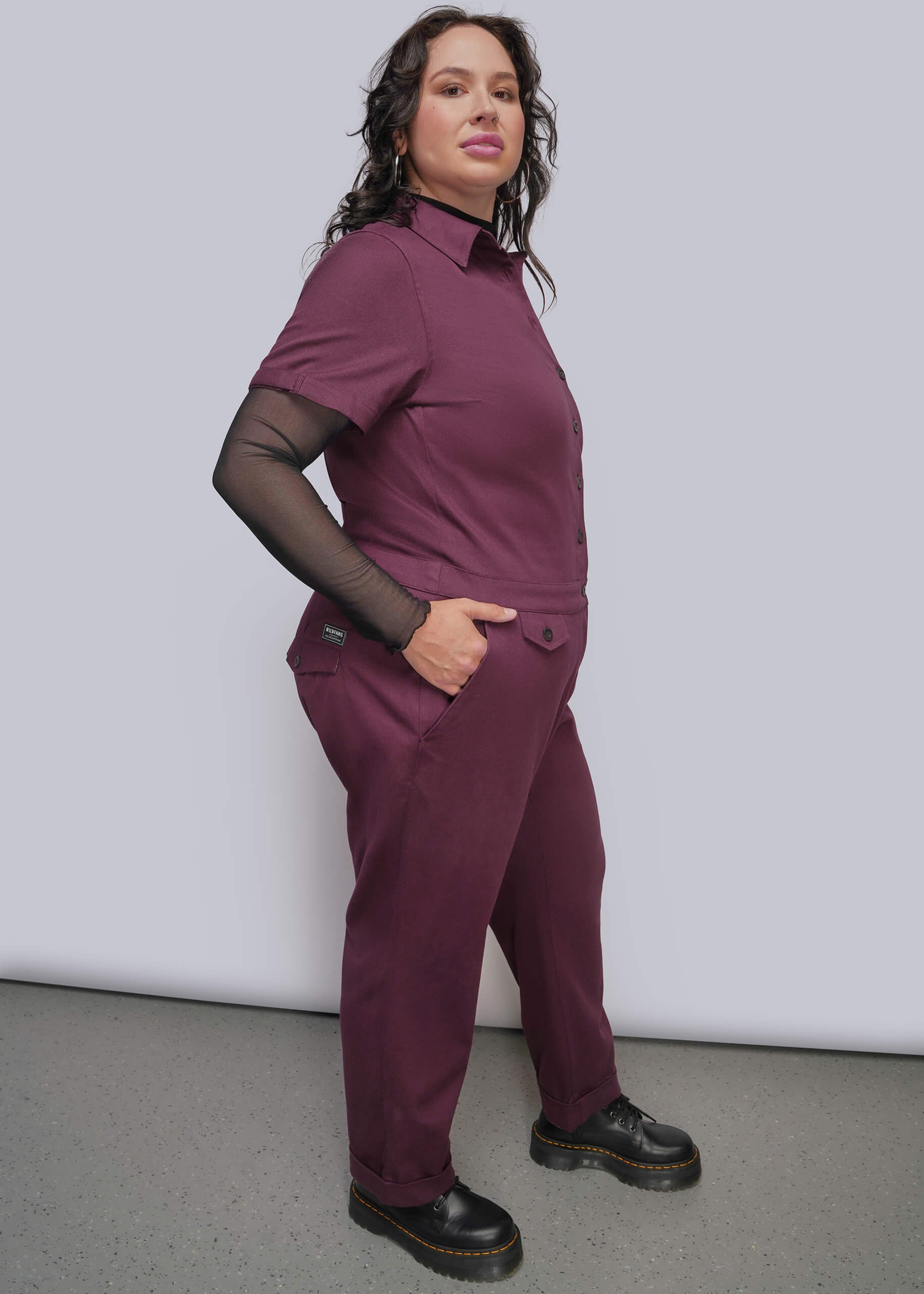 1X=A woman modeling a short sleeve burgundy jumpsuit with a sheer black long sleeve top underneath, paired with black boots, against a plain light gray studio background.