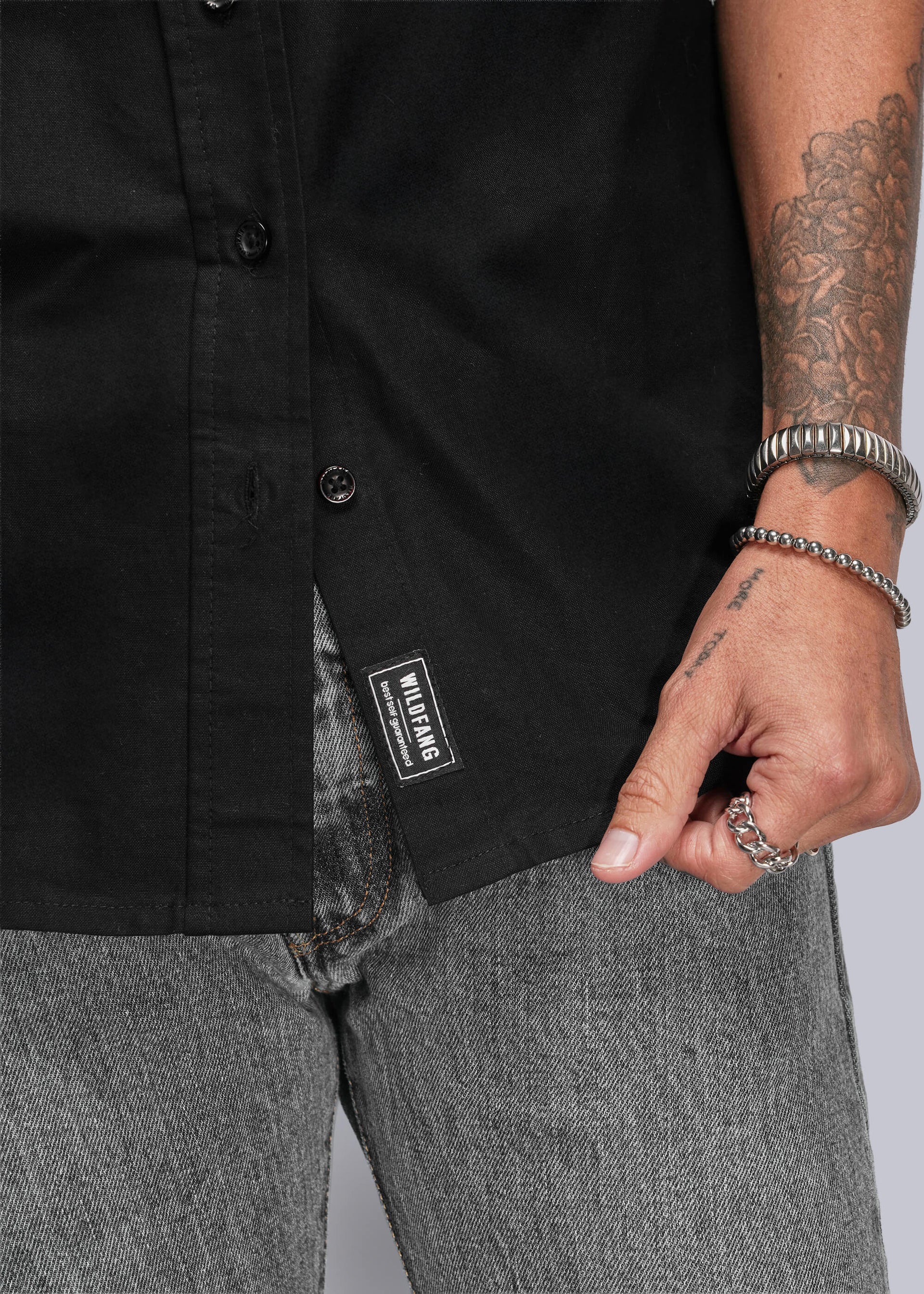S=Close up of person wearing a black shirt with a Wildfang label, gray pants, and multiple bracelets on a neutral background.