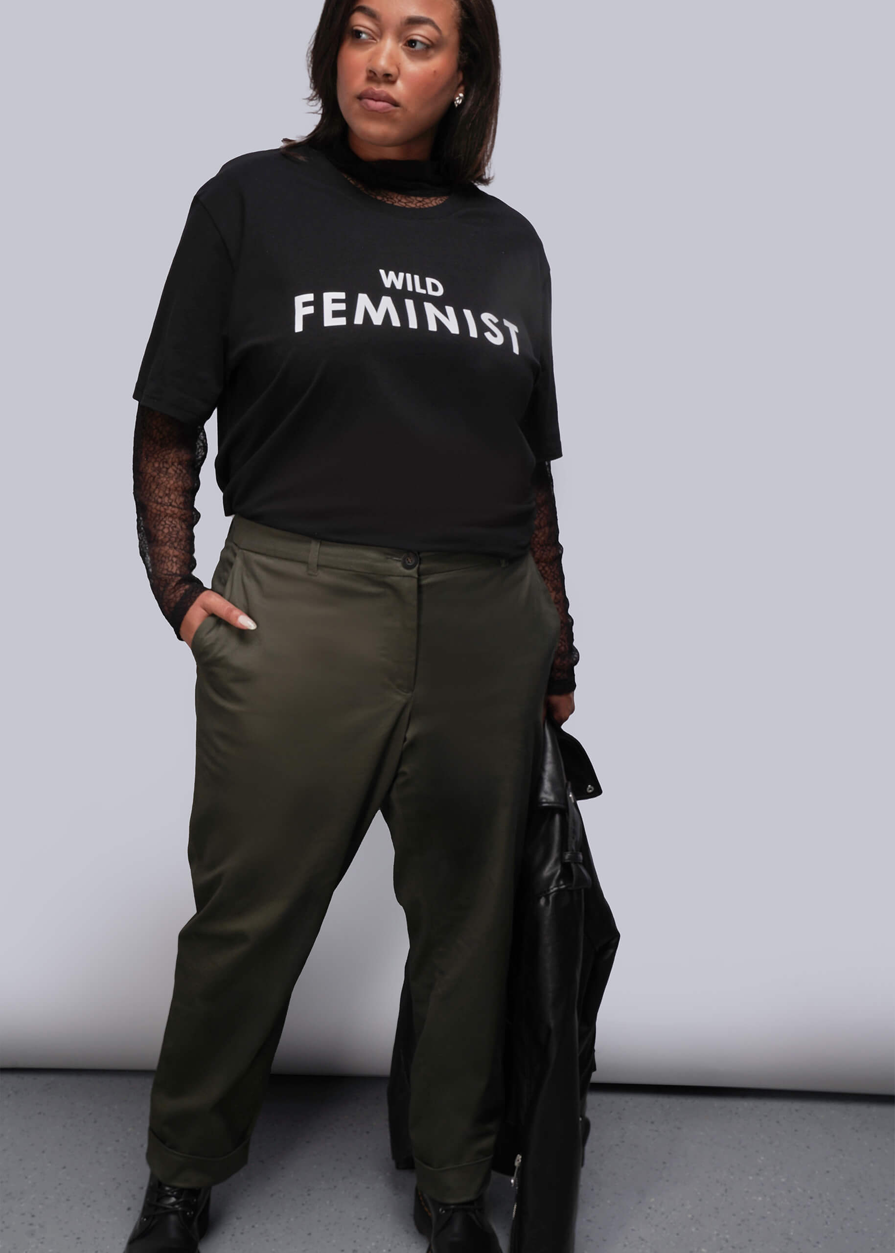 1X=Person wearing a black 'WILD FEMINIST' t-shirt and green pants against a plain background