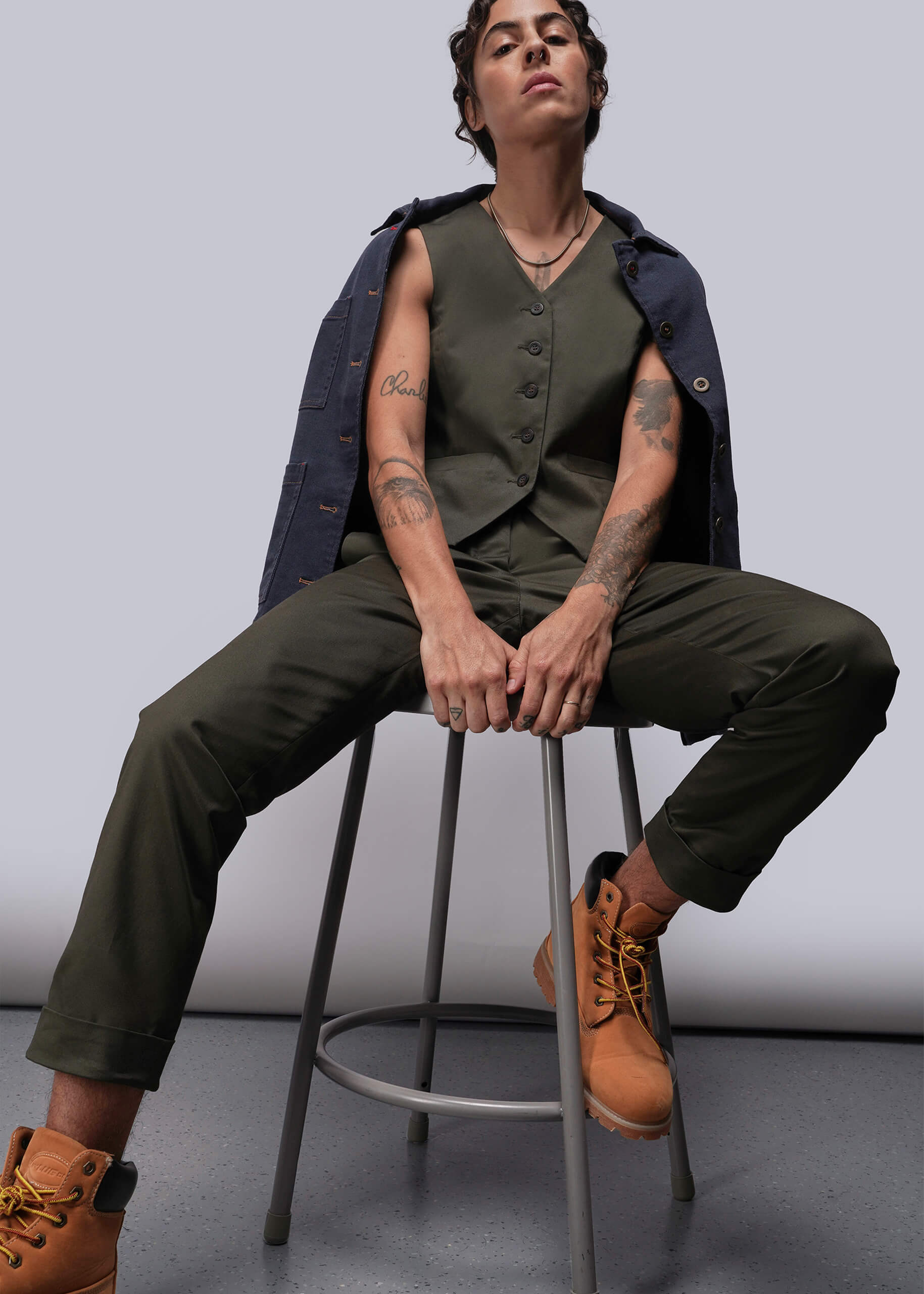 S=Person wearing a green vest and pants with brown boots sitting on a stool against a plain background
