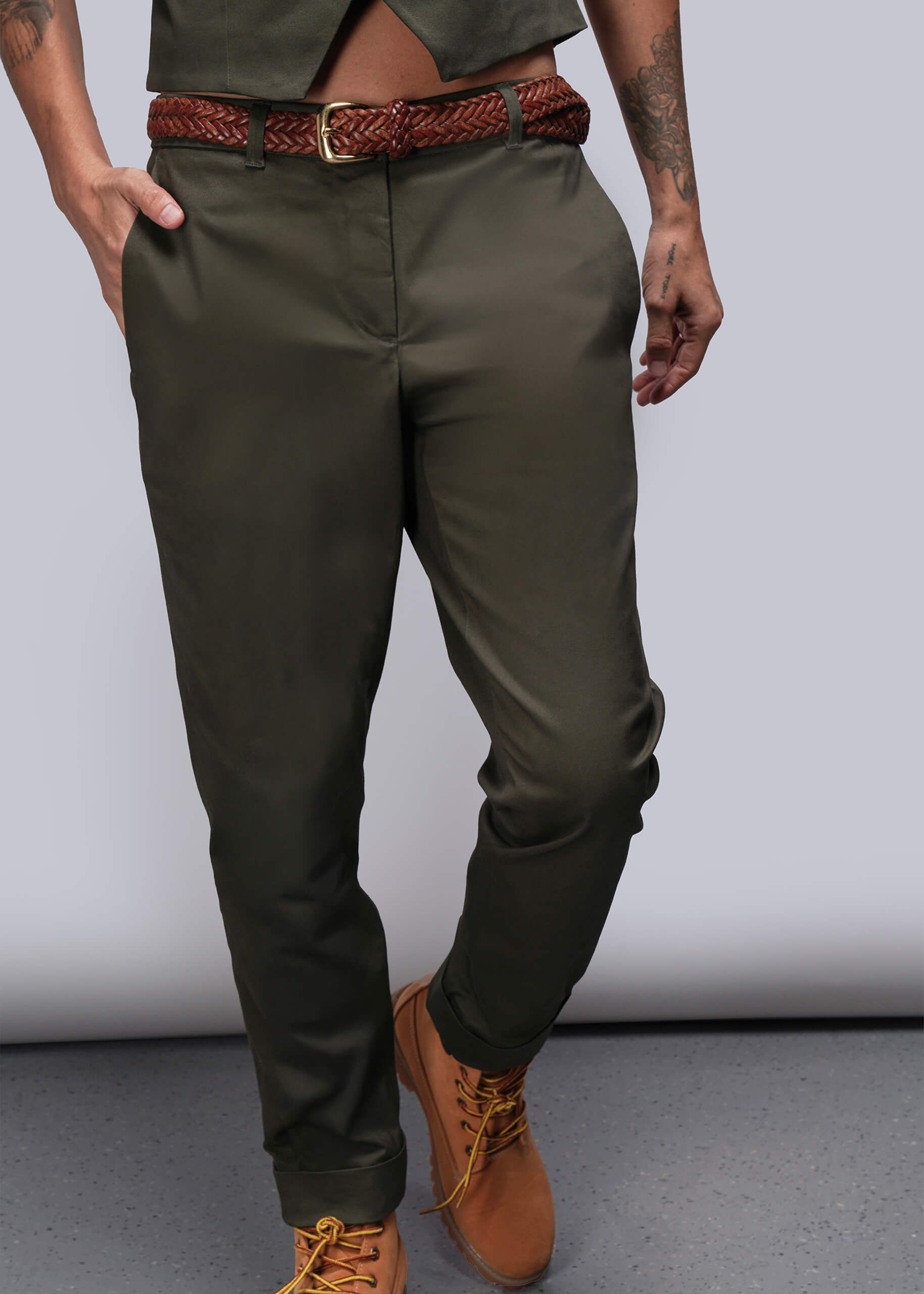 Person wearing olive green pants with a brown belt and shoes on a plain background